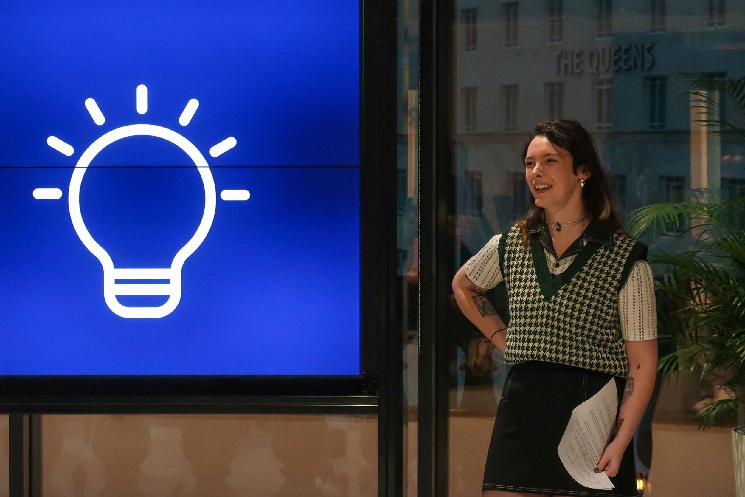 A woman stands near a large digital screen displaying a white light bulb icon on a blue background. She has dark hair, tattoos on her arms, and is smiling, holding some papers in her hand.