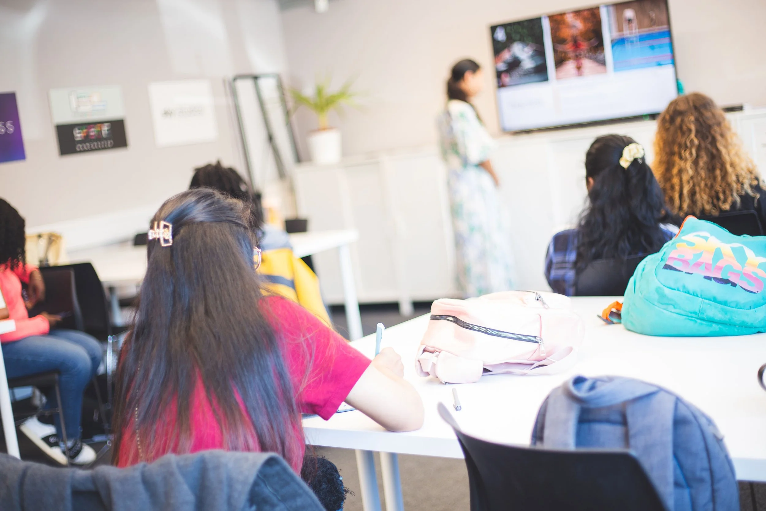 A classroom with students seated at desks, facing a teacher and a large screen displaying a presentation.