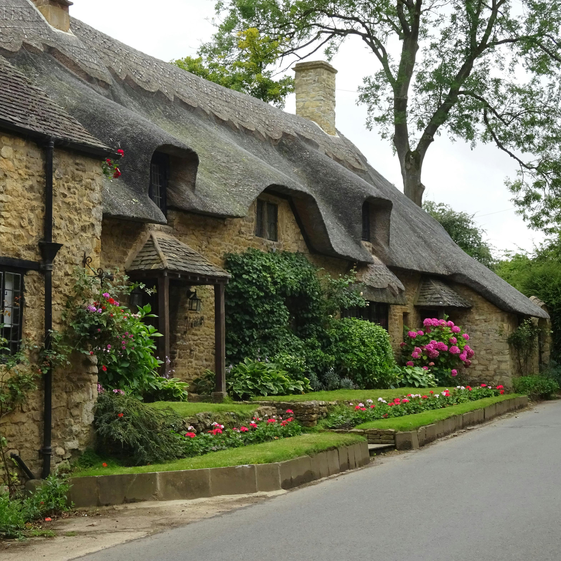 A quaint stone cottage with a thatched roof and colorful flowers in the garden along a paved street.