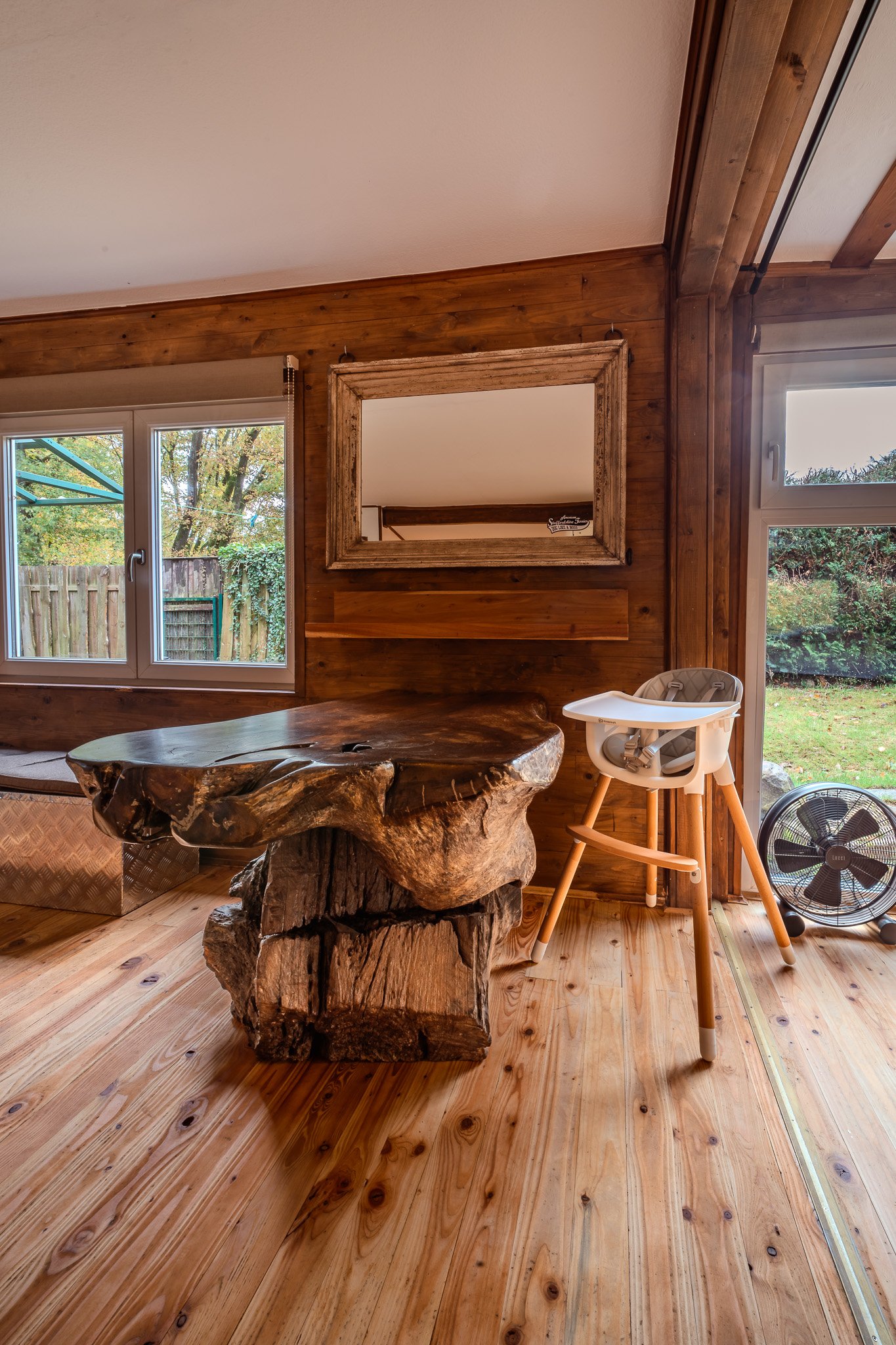 Ein rustikaler Raum mit Holzverkleidung, einem großen Baumstammtisch, einem weißen Hochstuhl, einem Wandspiegel und einem Ventilator, mit Blick auf einen Garten durch die Fenster.