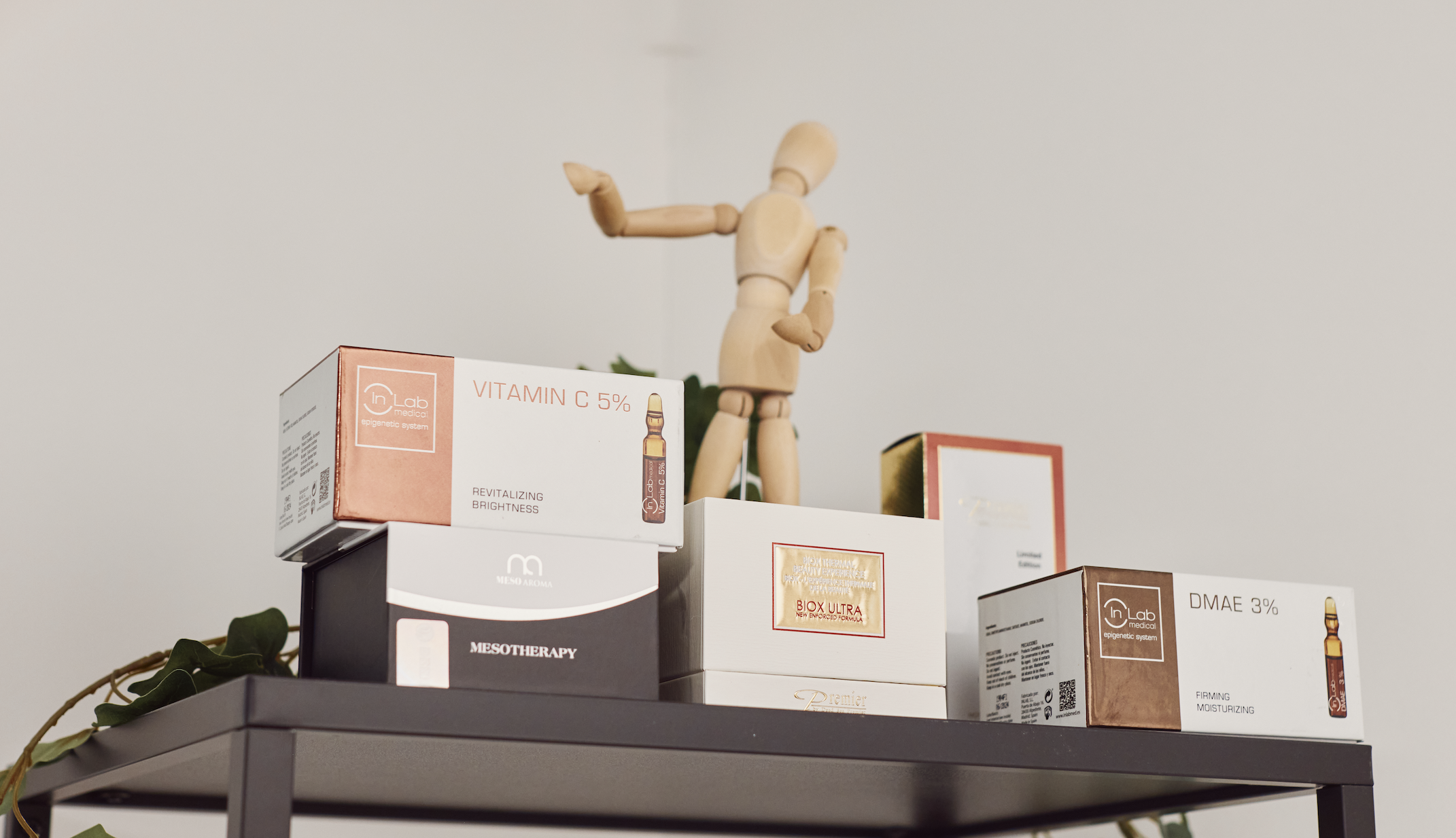 A shelf with boxes of skincare products, a wooden mannequin in a dancing pose, and some green leaves hanging over the edge.