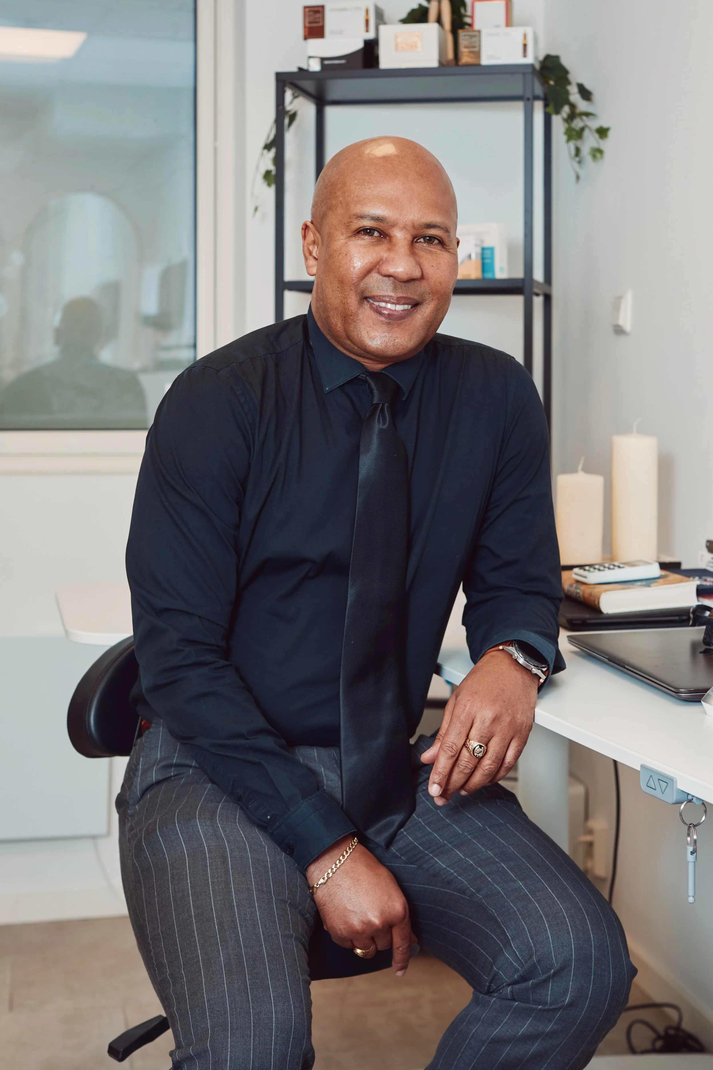 A man in a black shirt and tie sitting at a white desk in an office, smiling at the camera. There are candles, books, and a laptop on the desk behind him. Shelves with boxes and plants are visible in the background.