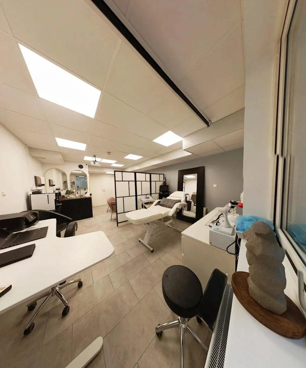 An interior room with medical or spa furnishings, including a treatment bed, a large mirror, a partition, a desk, chairs, and medical or skincare equipment.