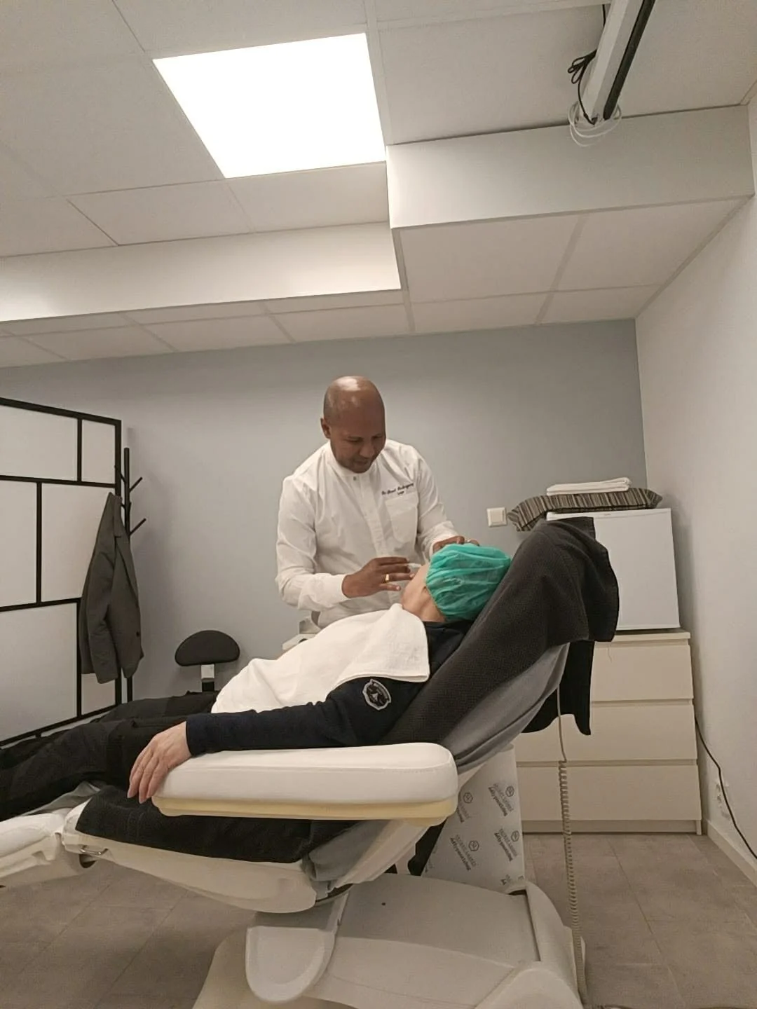 A person lying on a medical examination chair with a green surgical cap, while a healthcare professional in white scrubs examines their face in a clinic or hospital room.