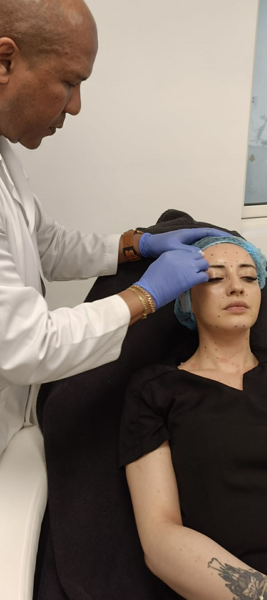 A woman receiving an injection in her forehead from a healthcare professional wearing blue gloves, a white coat, and a gold bracelet, in a clinical setting.