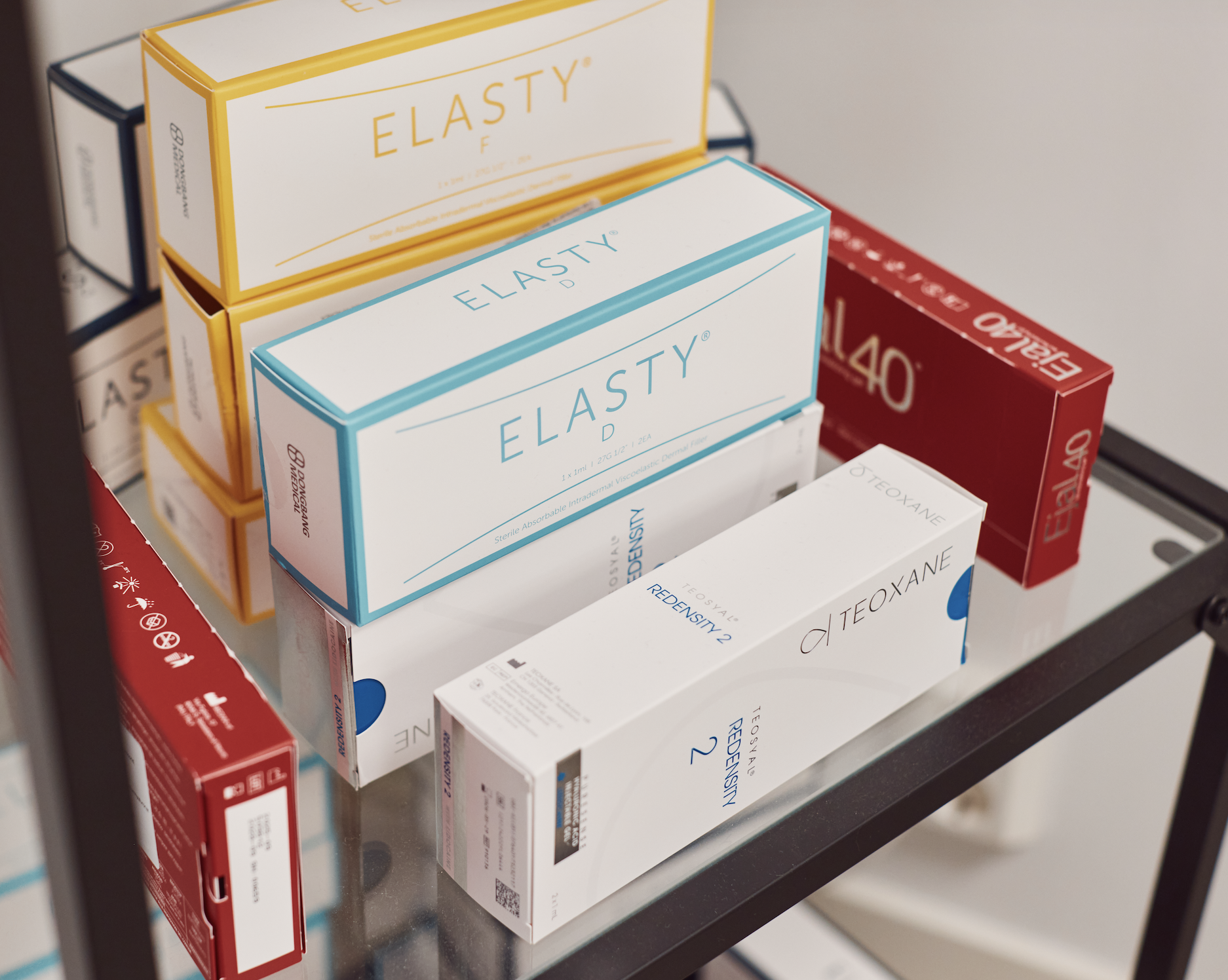 Various boxes of dermal fillers on a glass shelf, including ElastY, Teoxane, and Belotero brand products.