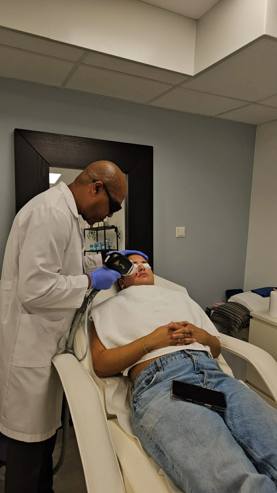 A medical professional using a handheld device on a woman lying on a hospital bed, with a smartphone placed on her stomach, in a clinical setting.