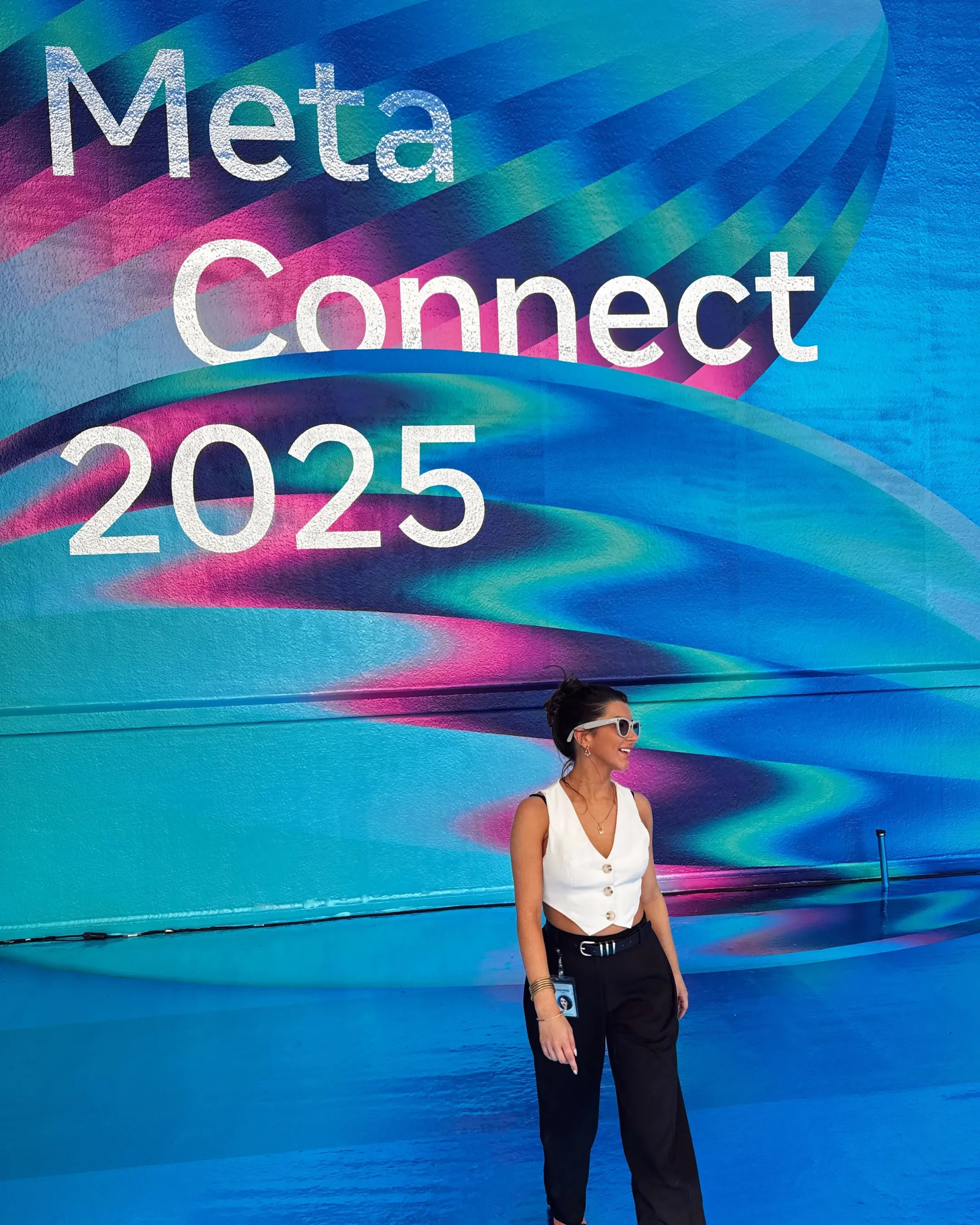 A woman with dark hair tied back, wearing white sunglasses, a white sleeveless top, and black pants with a badge hanging from her waistband, standing in front of a colorful mural with the words 'Meta Connect 2025'.