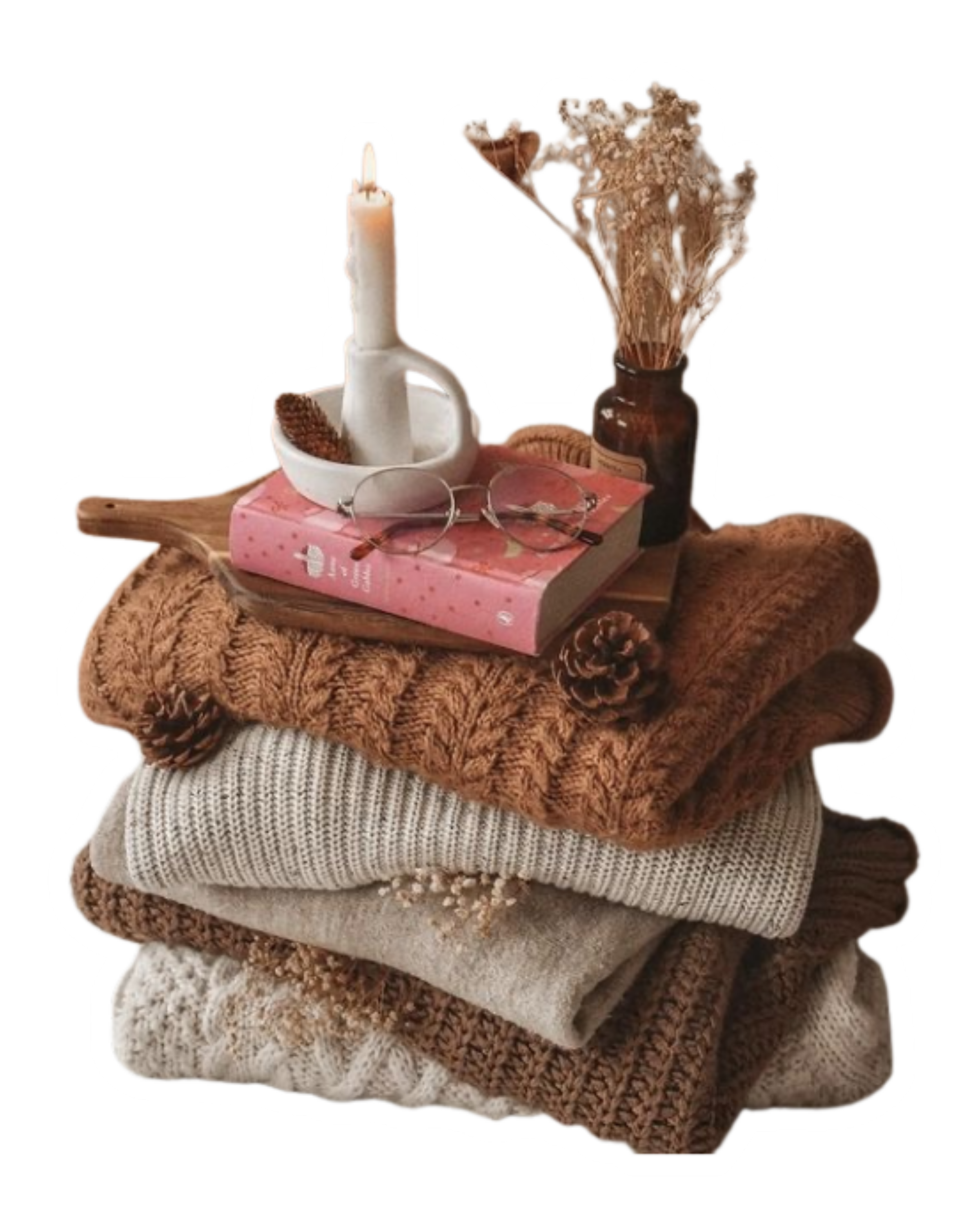 Stack of folded cozy sweaters with a wooden tray on top holding a pink book, a pair of eyeglasses, a white candle in a holder, a small white dish, pine cones, a dried flower arrangement in a brown bottle, and a cookie.