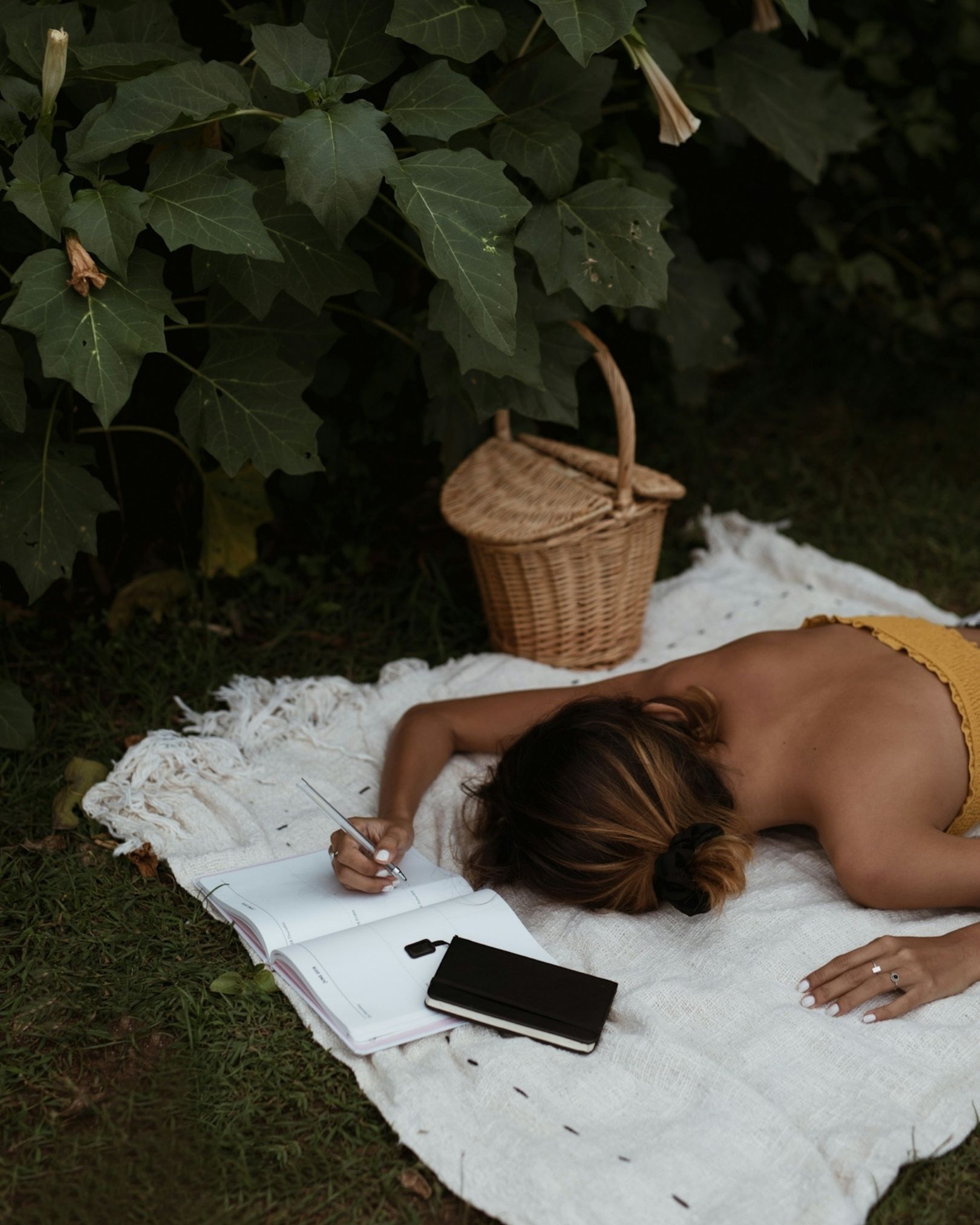 Woman lying on a white blanket outdoors, writing in a notebook with a pen, surrounded by green bushes, with a small woven basket nearby.
