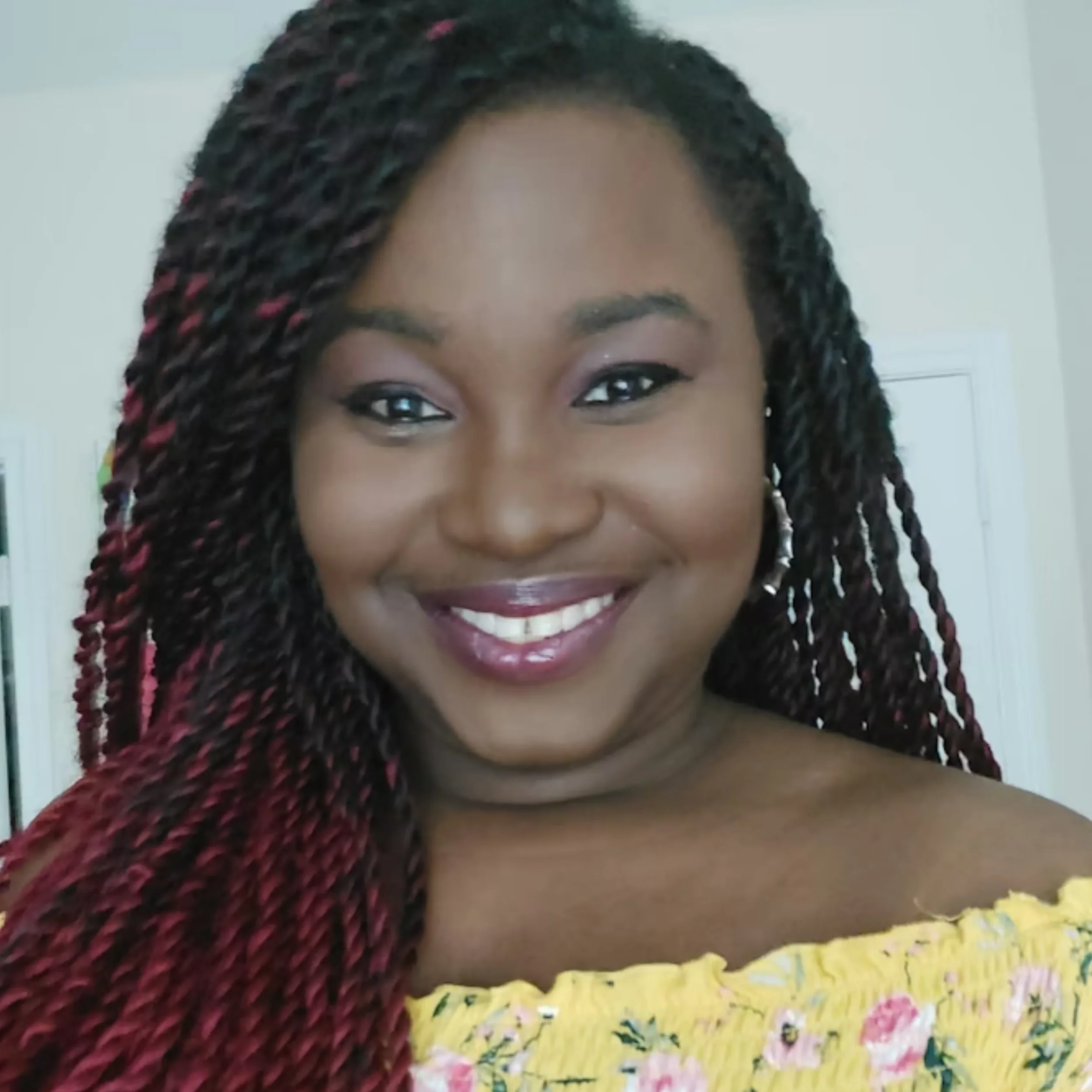 Close-up of a smiling woman with braided hair, wearing a yellow floral dress, indoors in front of a white wall.