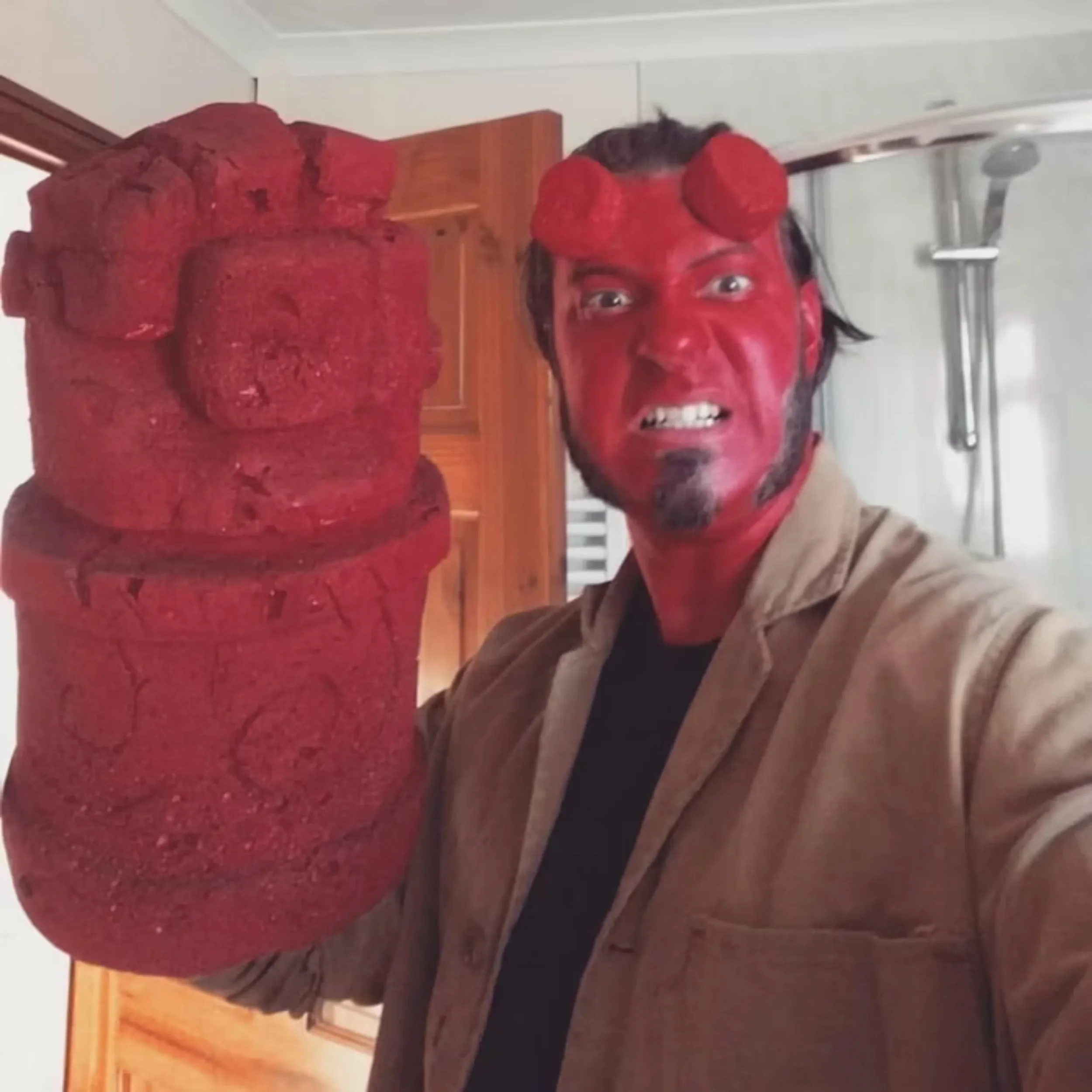 A person with red face and hair, making an angry facial expression, taking a selfie in front of a carved wooden statue of a person's head.