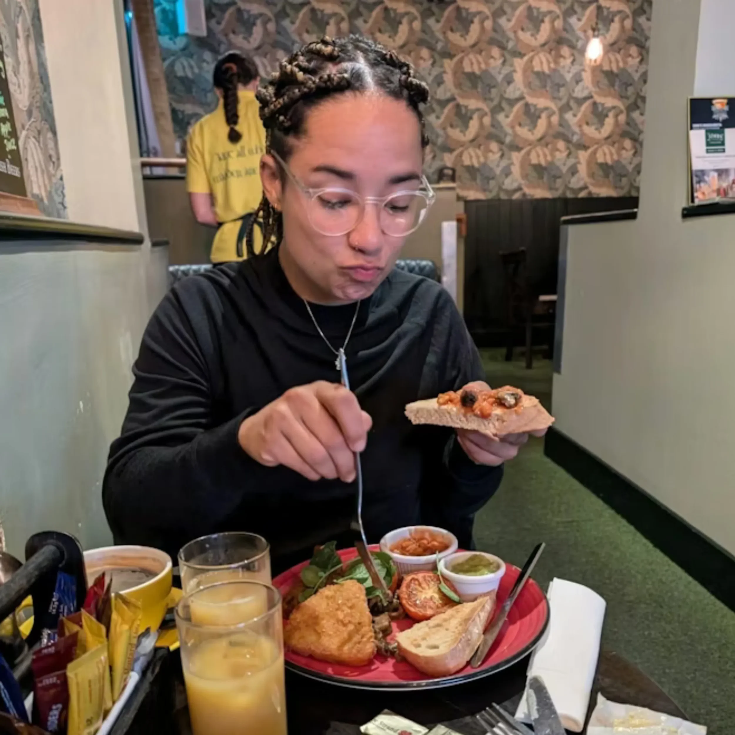 A woman with braided hair and glasses eating pizza at a restaurant table. The table has various foods including fried chicken, bread, and dips, along with beverages like juice. In the background, a person in a yellow shirt is visible.