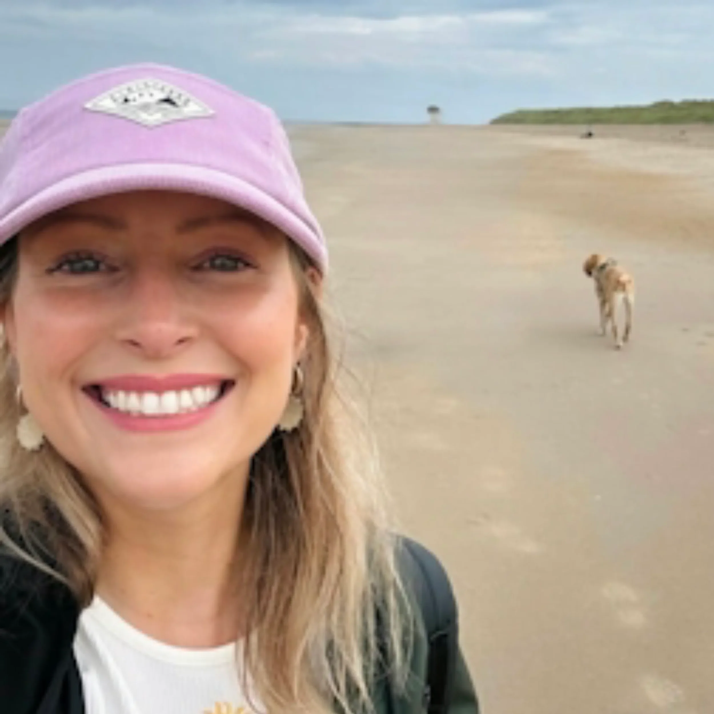 A person wearing a pink cap with a white patch on the front, smiling, standing on the beach with a dog in the background.