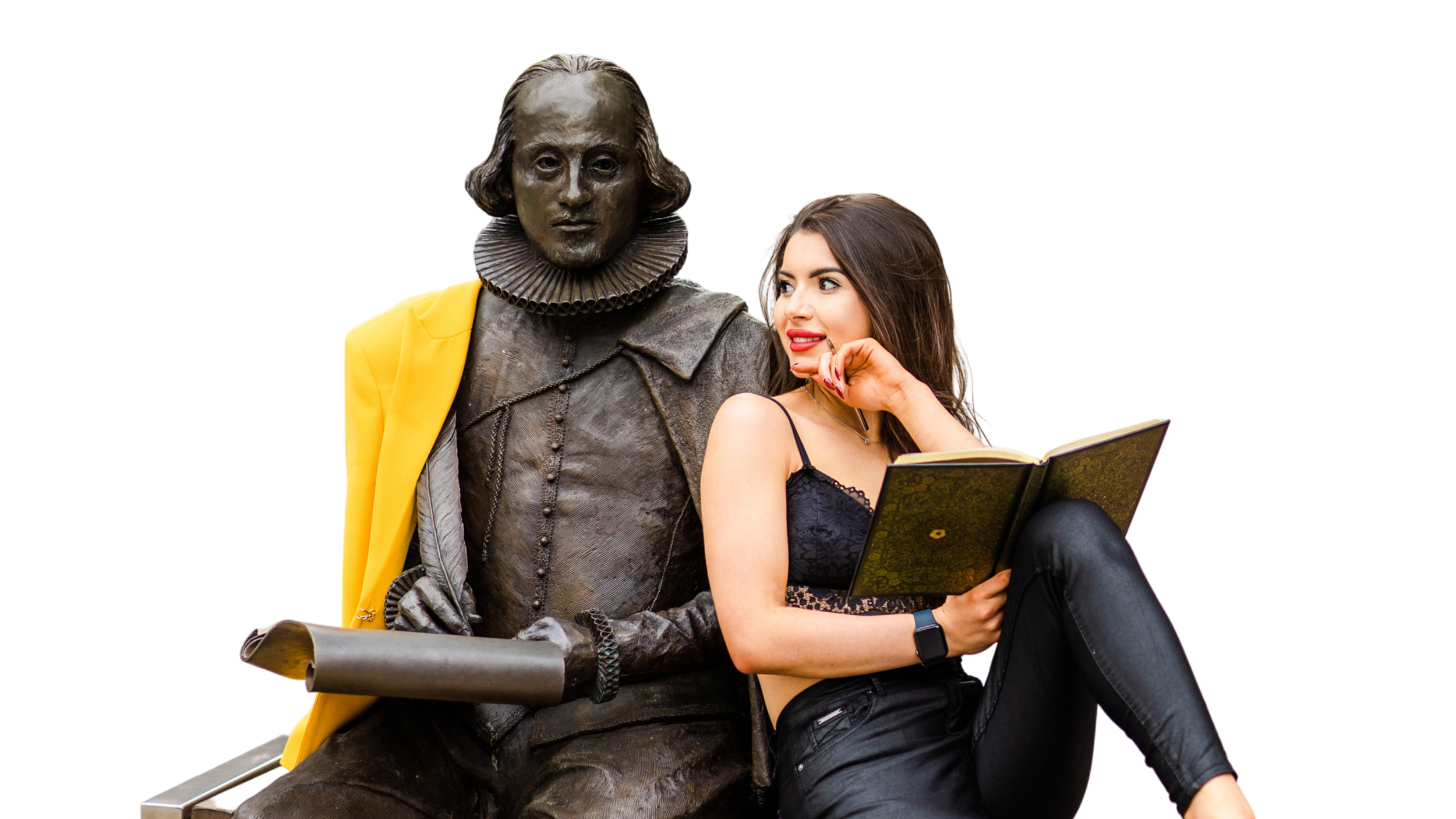 A young woman with long brown hair, wearing a black lace top and black leather pants, sitting next to a statue of William Shakespeare on a bench. She is holding an open book and appears to be talking or smiling at the statue.