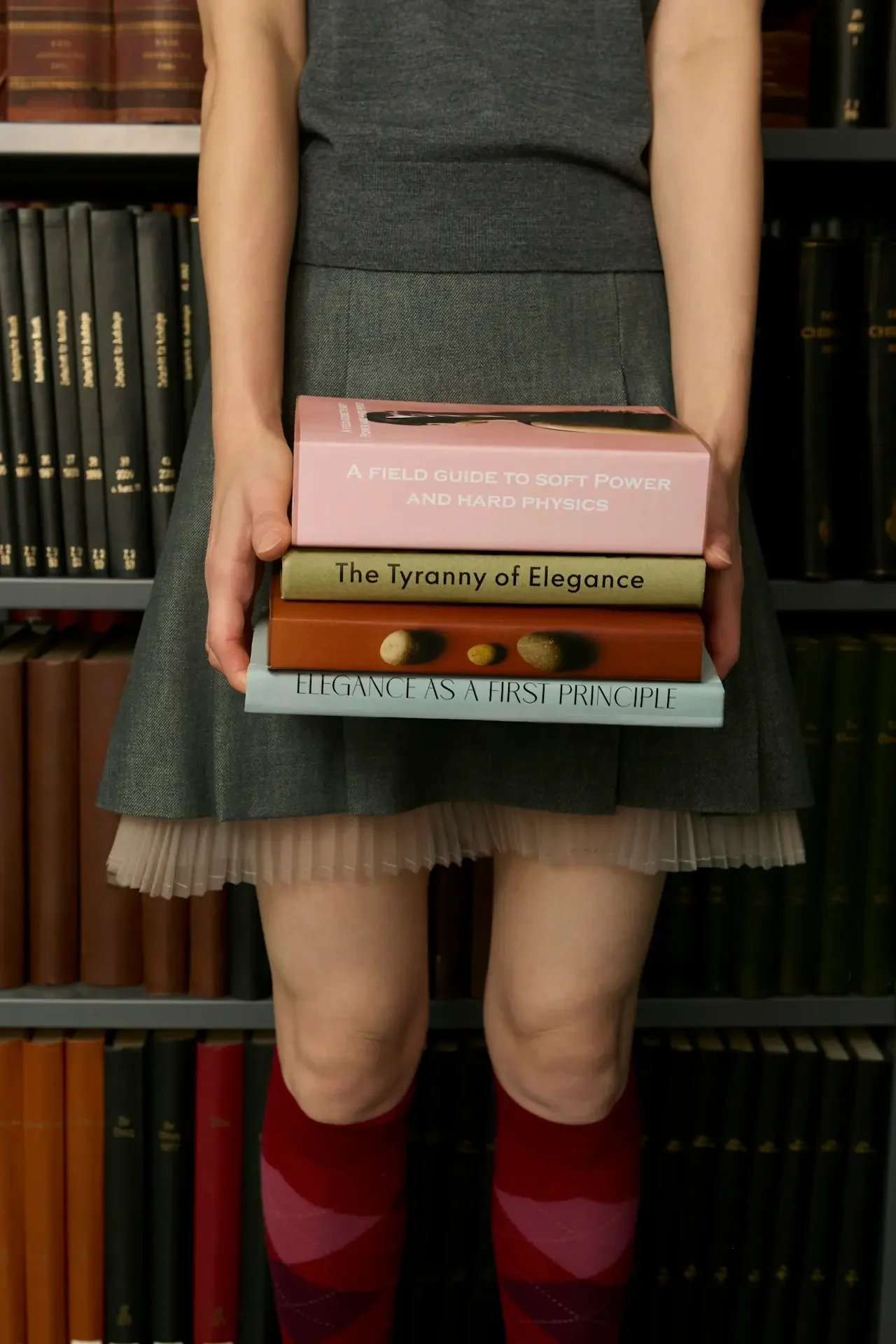 A person in a gray skirt and red argyle socks holds four books, with bookshelves in the background.