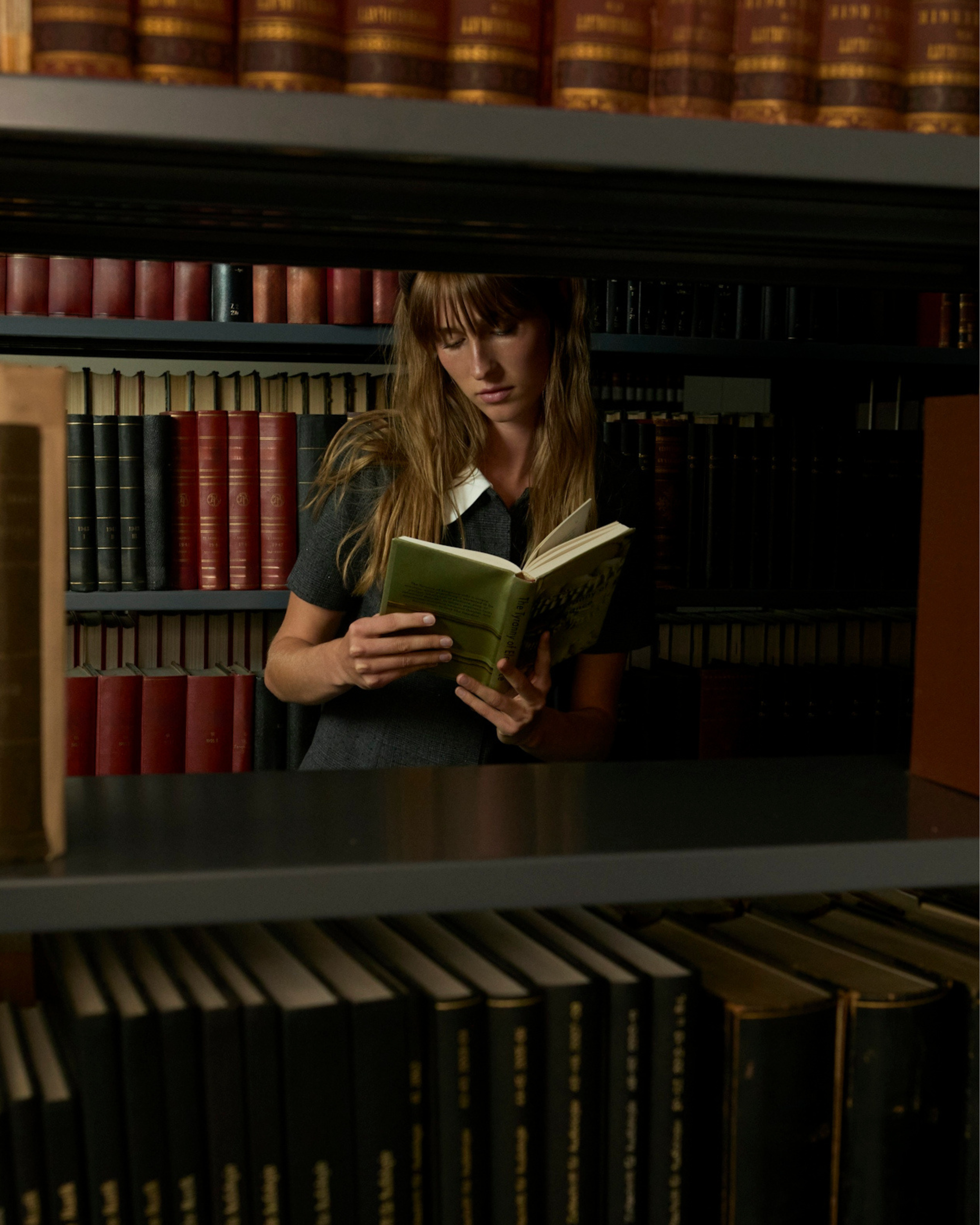 A woman with long brown hair, wearing a dark dress with a white collar, is reading a book in a dimly lit library between bookshelves filled with large books.