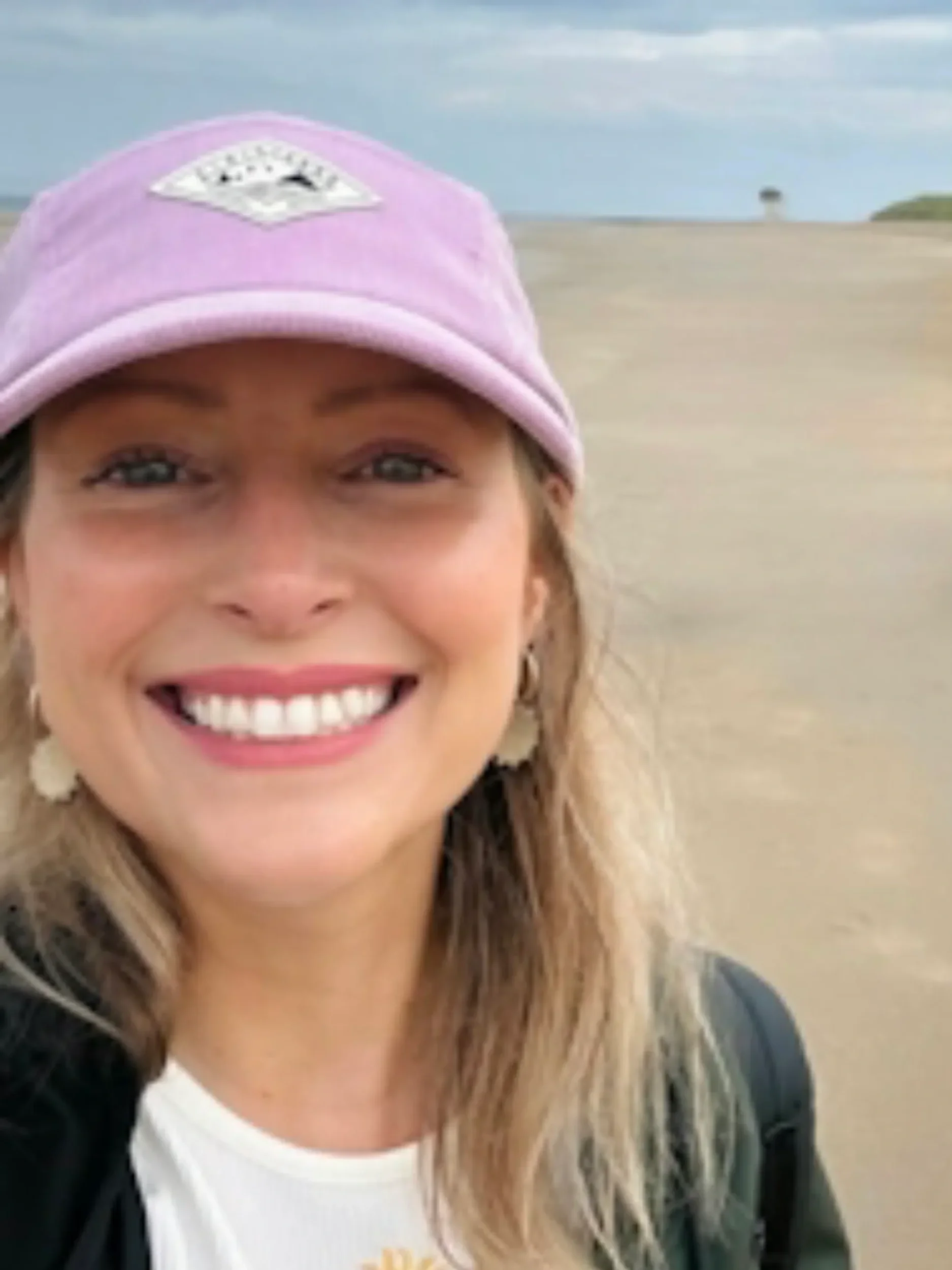 Close-up of a woman's smiling face with light brown hair, wearing a pink cap with a white patch, and a white shirt, outdoors on a sandy beach with a cloudy sky in the background.