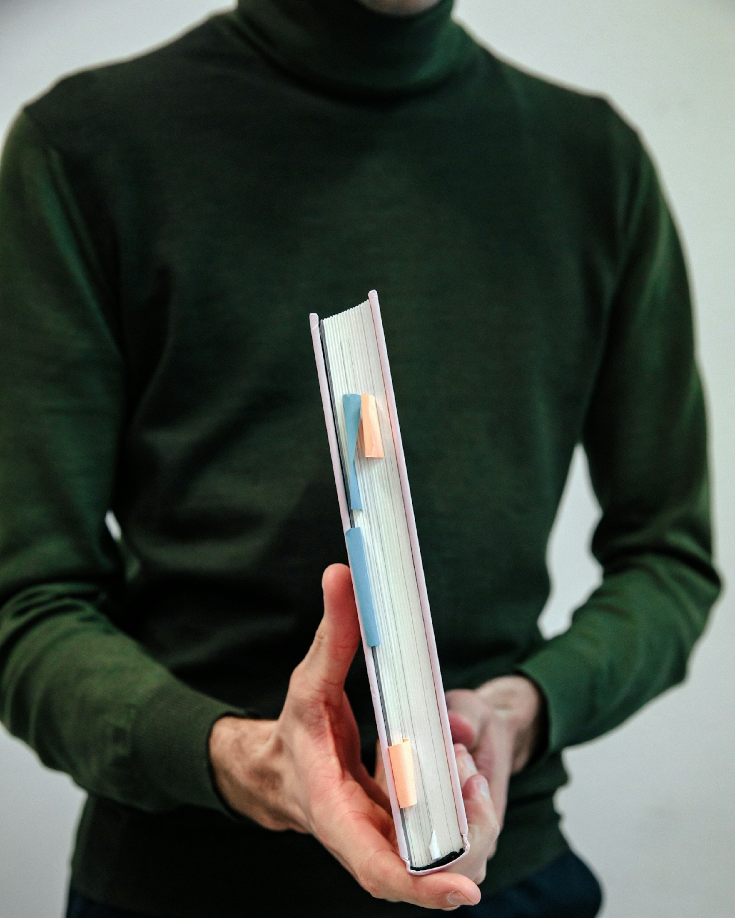 Person in a green turtleneck holding a hardcover book with sticky notes in the pages.