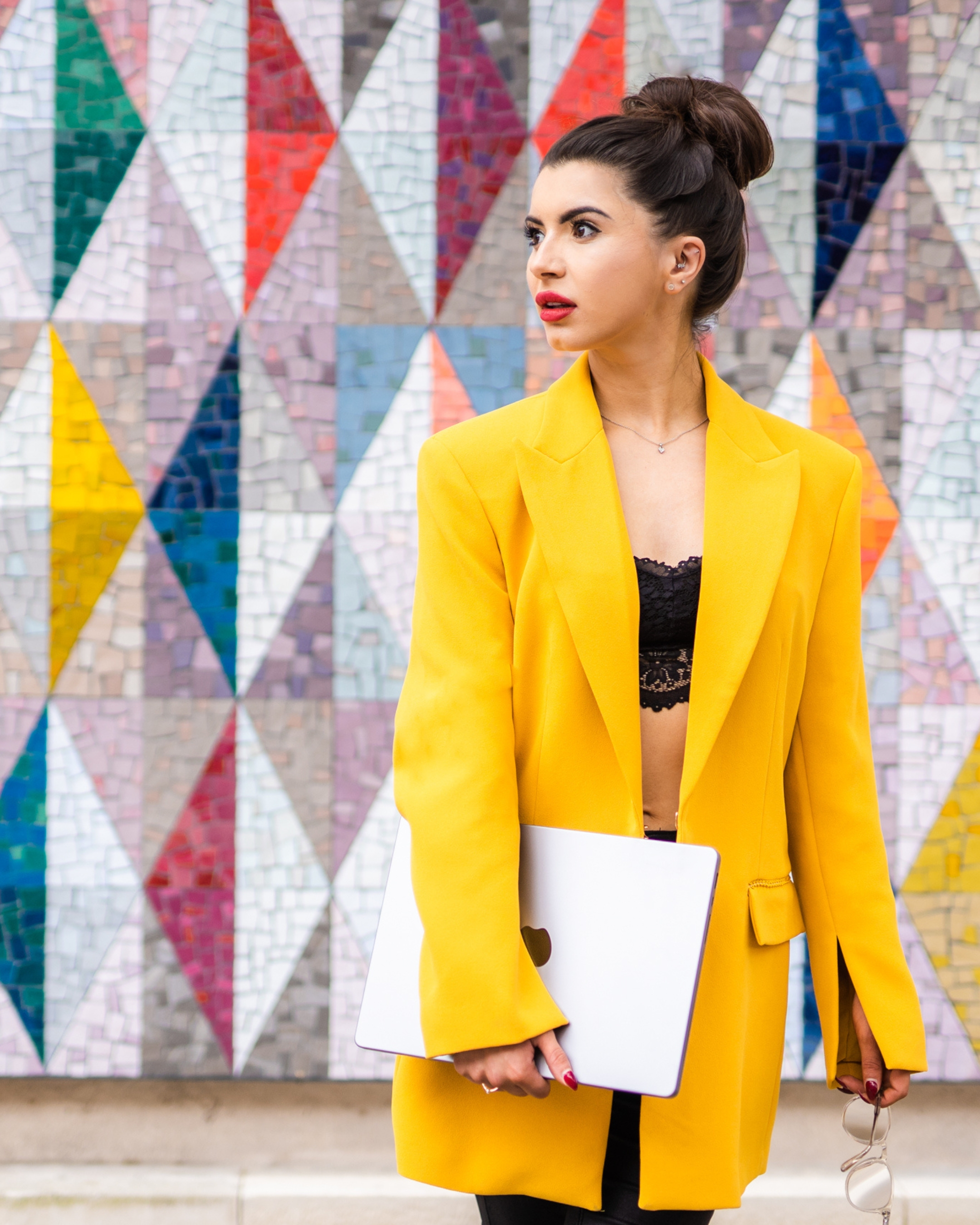 A woman with dark hair styled in a bun wearing a yellow blazer, holding a closed laptop, standing in front of a colorful mosaic wall.