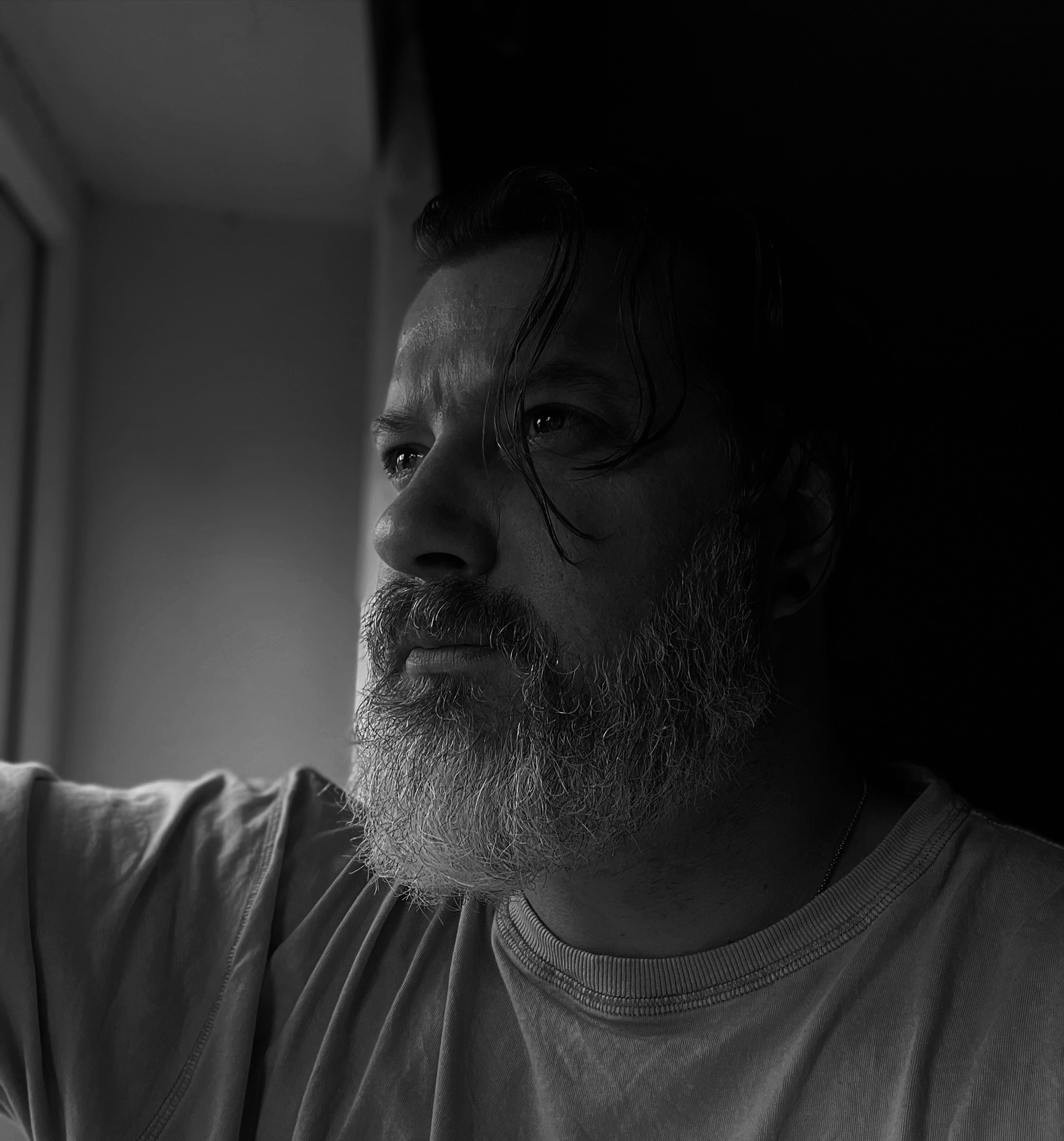 A man with a beard and long dark hair looking out a window, wearing a striped shirt and a grey jacket, in black and white.