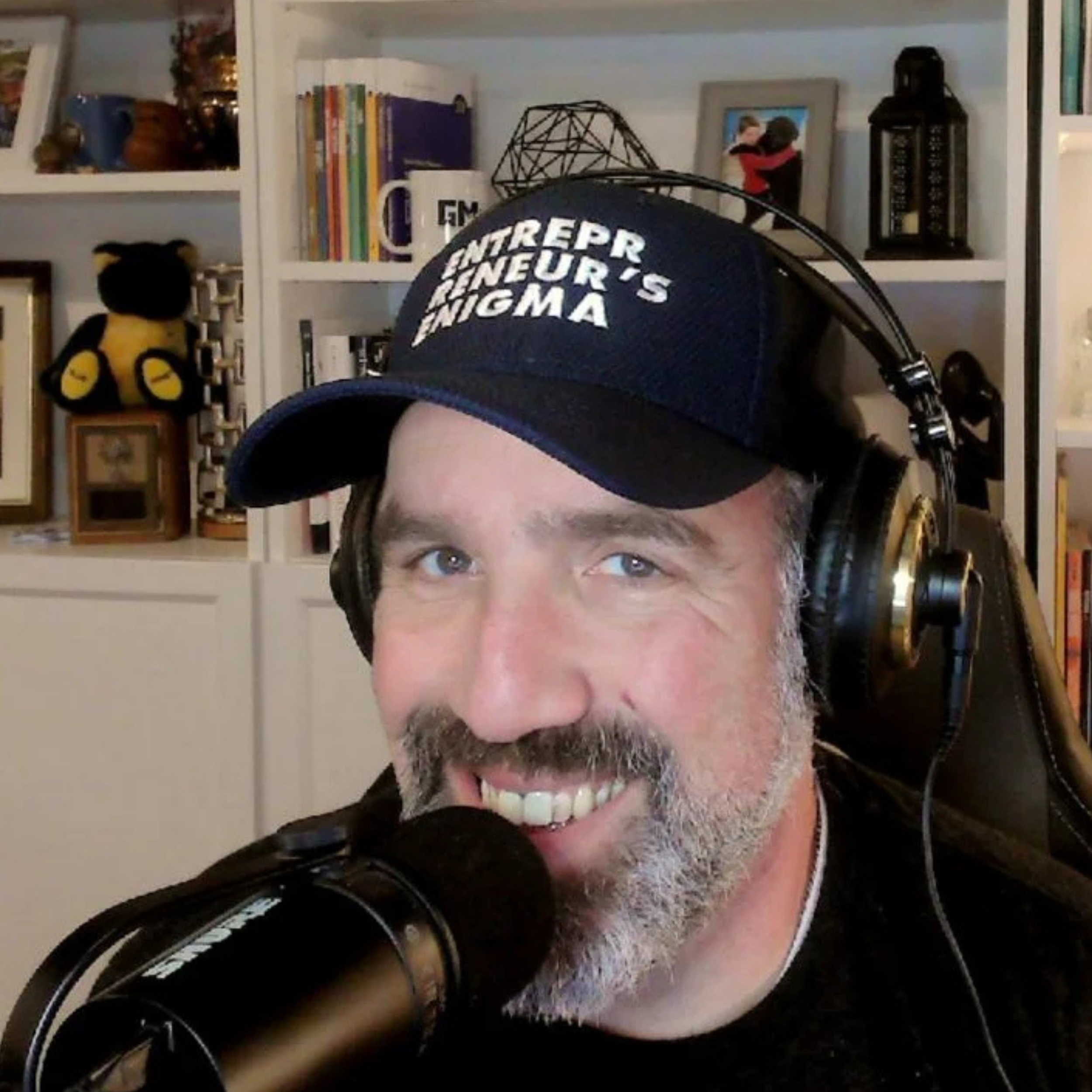 A man with a beard and mustache wearing a black cap with white text, large headphones, and speaking into a microphone, sitting in front of a bookshelf decorated with various framed photos, books, and objects.