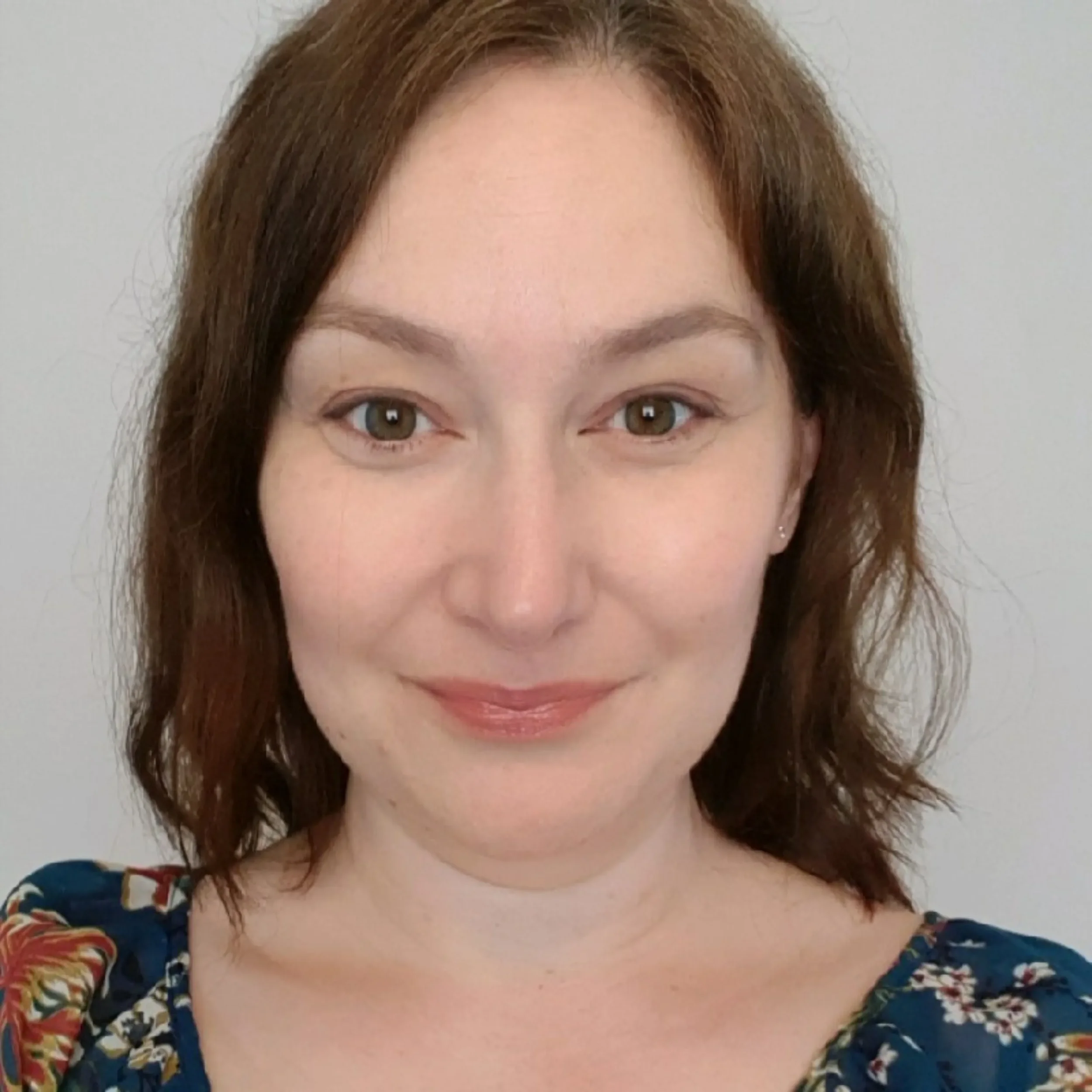 Close-up of a woman with shoulder-length brown hair, hazel eyes, light makeup, and wearing a floral top, smiling at the camera.