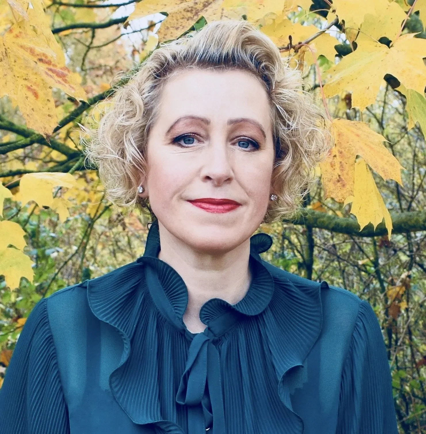 A woman with short, curly blonde hair and blue eyes, wearing a navy blue blouse with ruffles and a bow at the collar, standing outdoors in front of yellow autumn leaves.