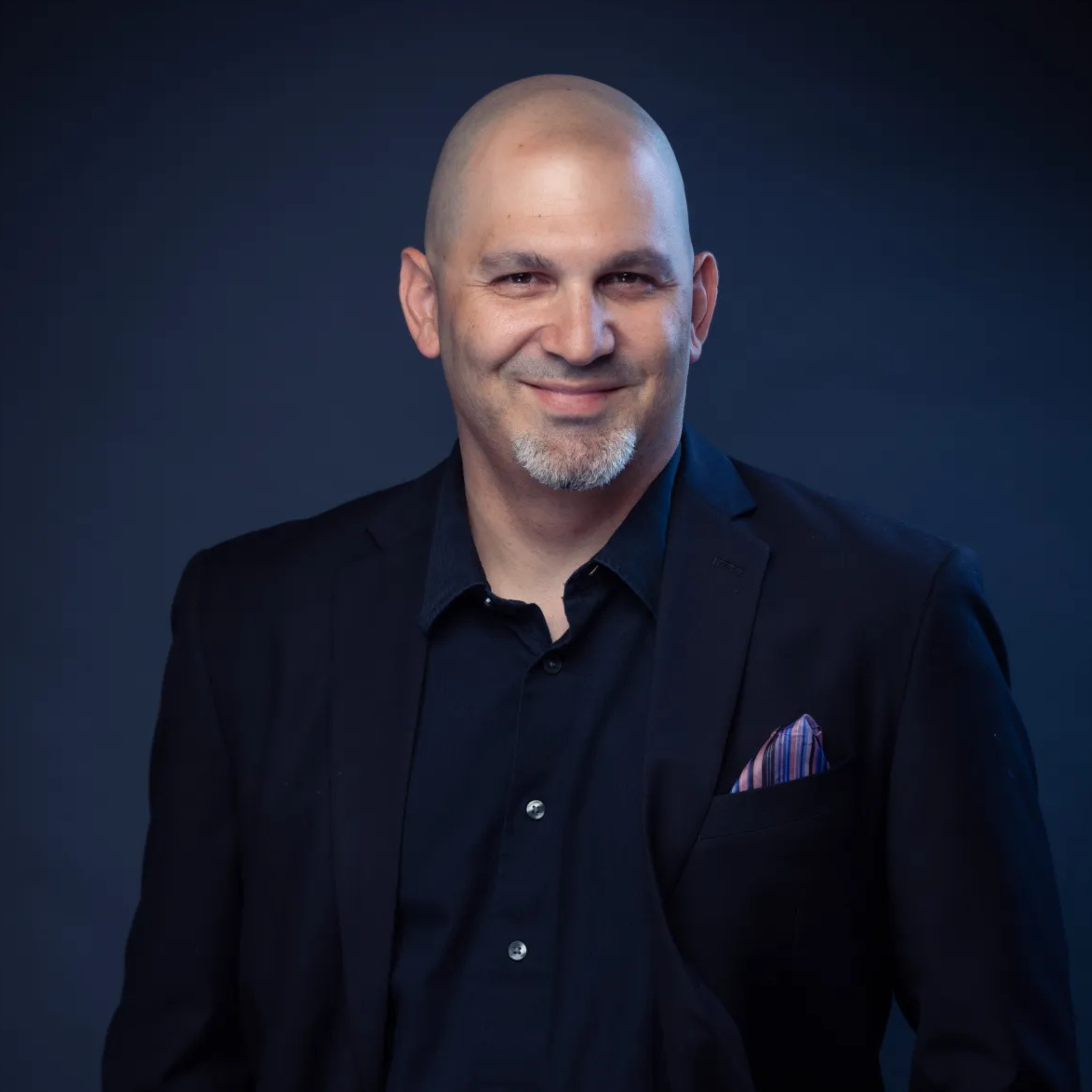 Headshot of a smiling man with a shaved head and beard, wearing a dark suit and shirt, against a dark background.
