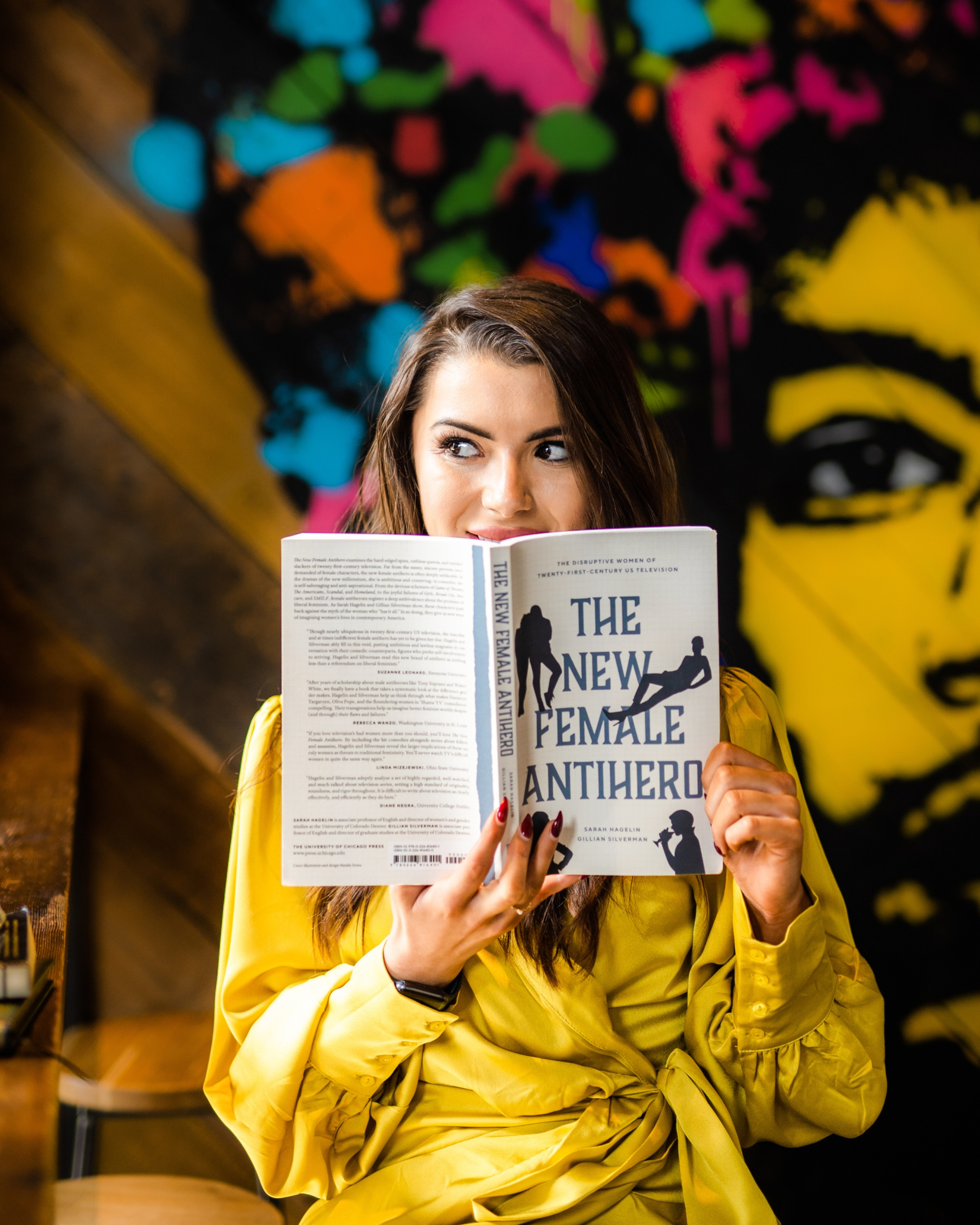 A woman in a yellow jacket with long brown hair holds an open book that partially obscures her face, showing her eyes and forehead. She is in a room with a colorful graffiti-style mural on the wall behind her, featuring vibrant shades of pink, orange, blue, green, and yellow.