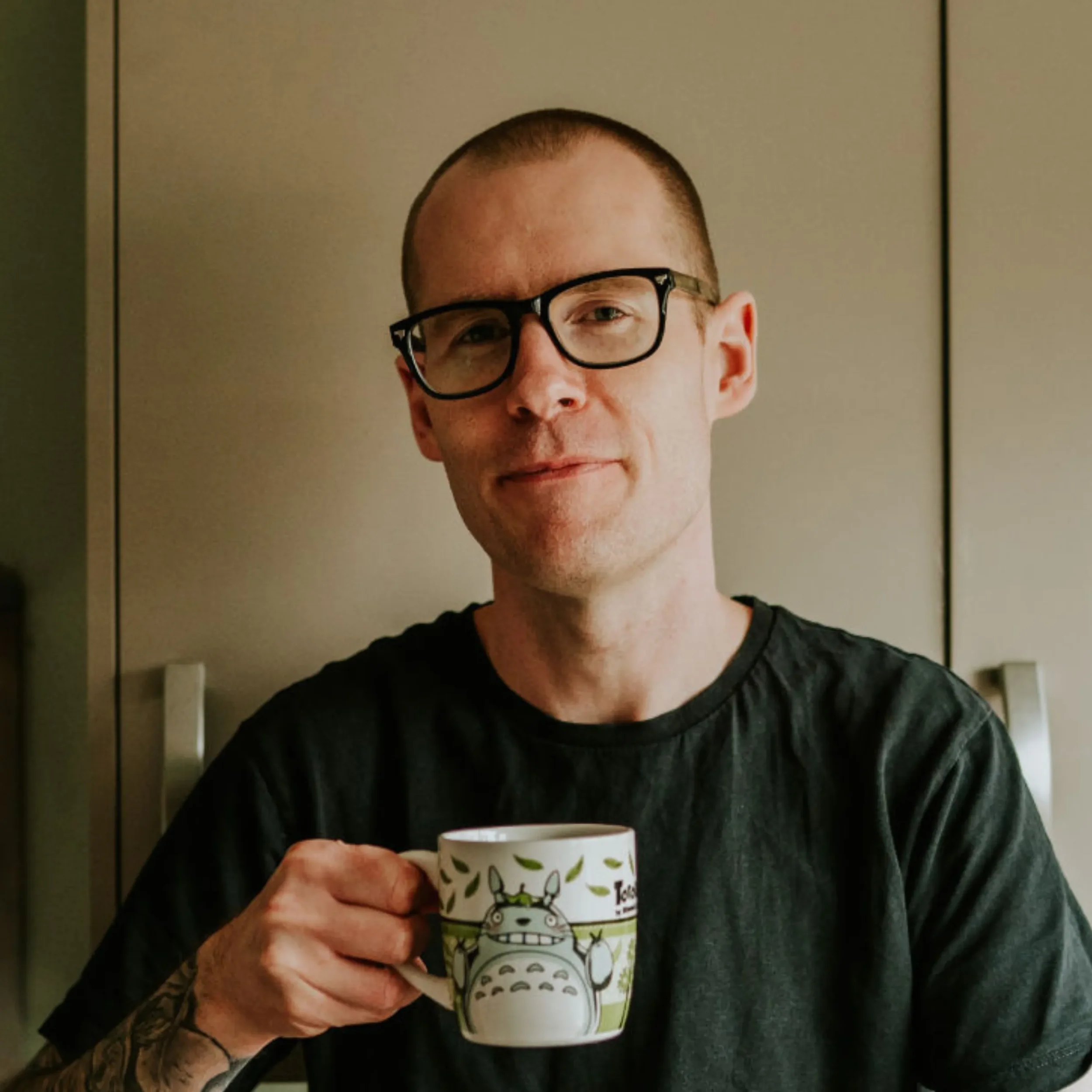 A man with glasses and a shaved head holding a Totoro-themed mug, standing in front of a plain background.