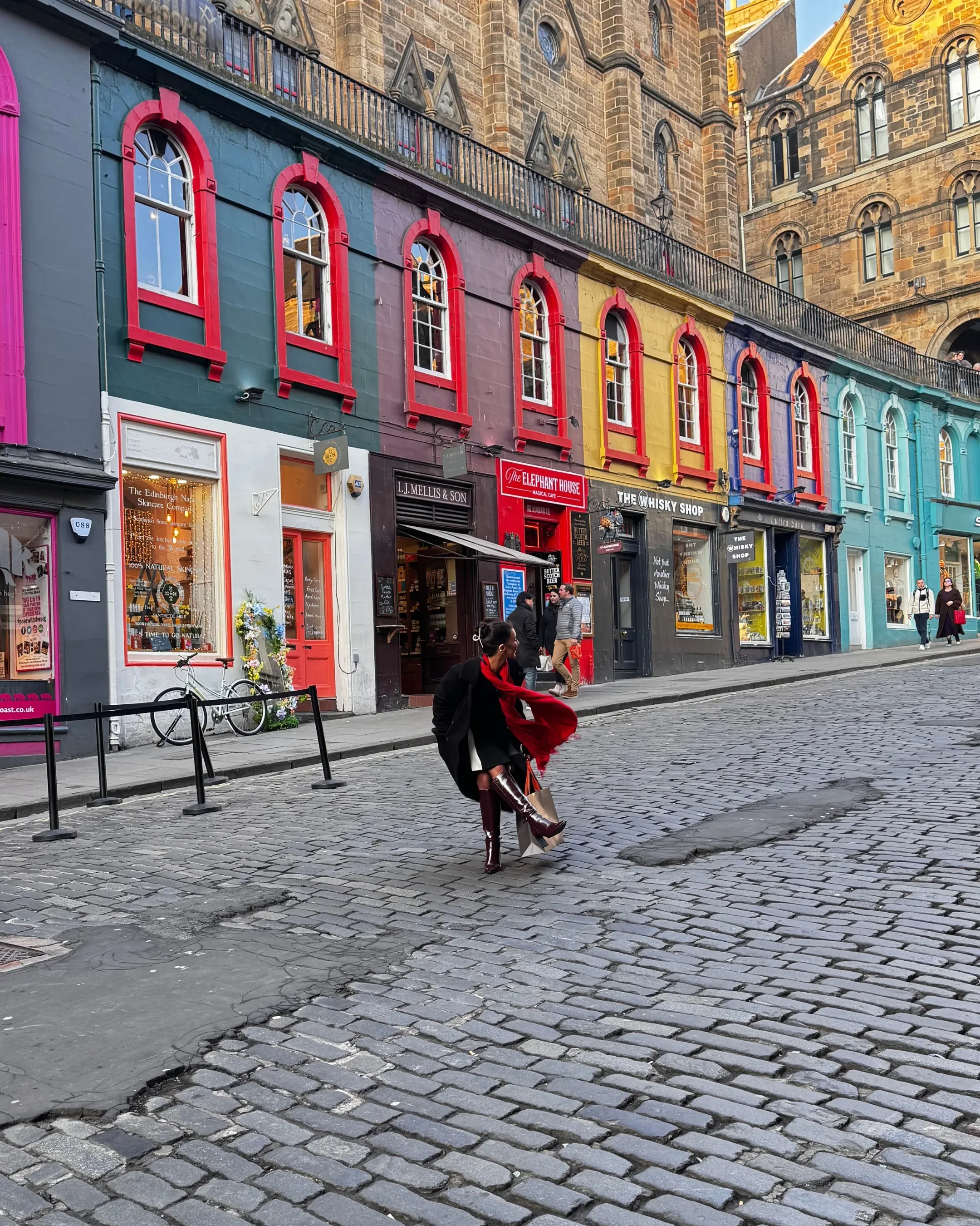 Colorful row of shops on a cobblestone street with people walking, in front of a historic stone building.