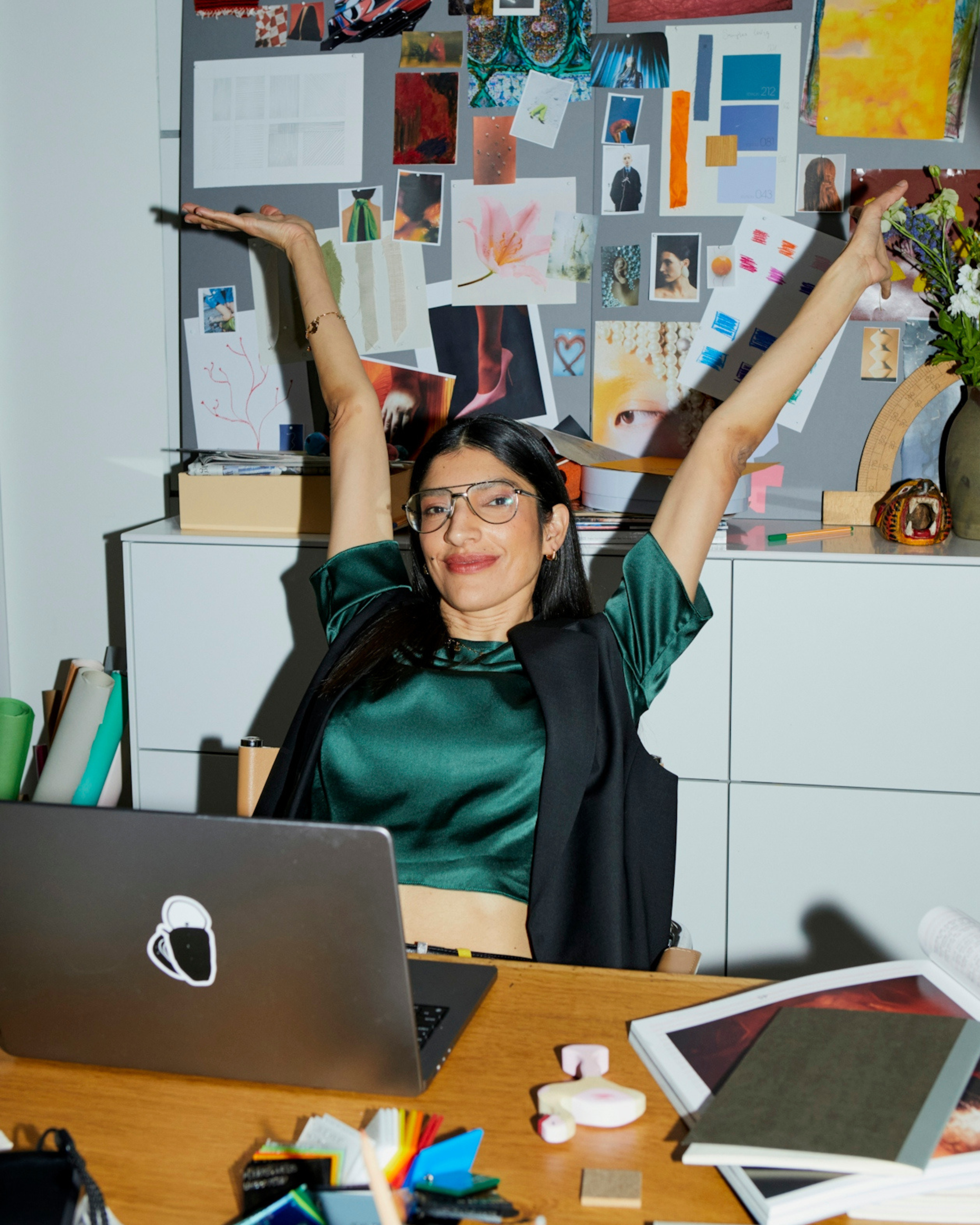 A woman with glasses sitting at a desk with her arms raised, smiling, in front of a cluttered workspace and a bulletin board covered with colorful images and notes.