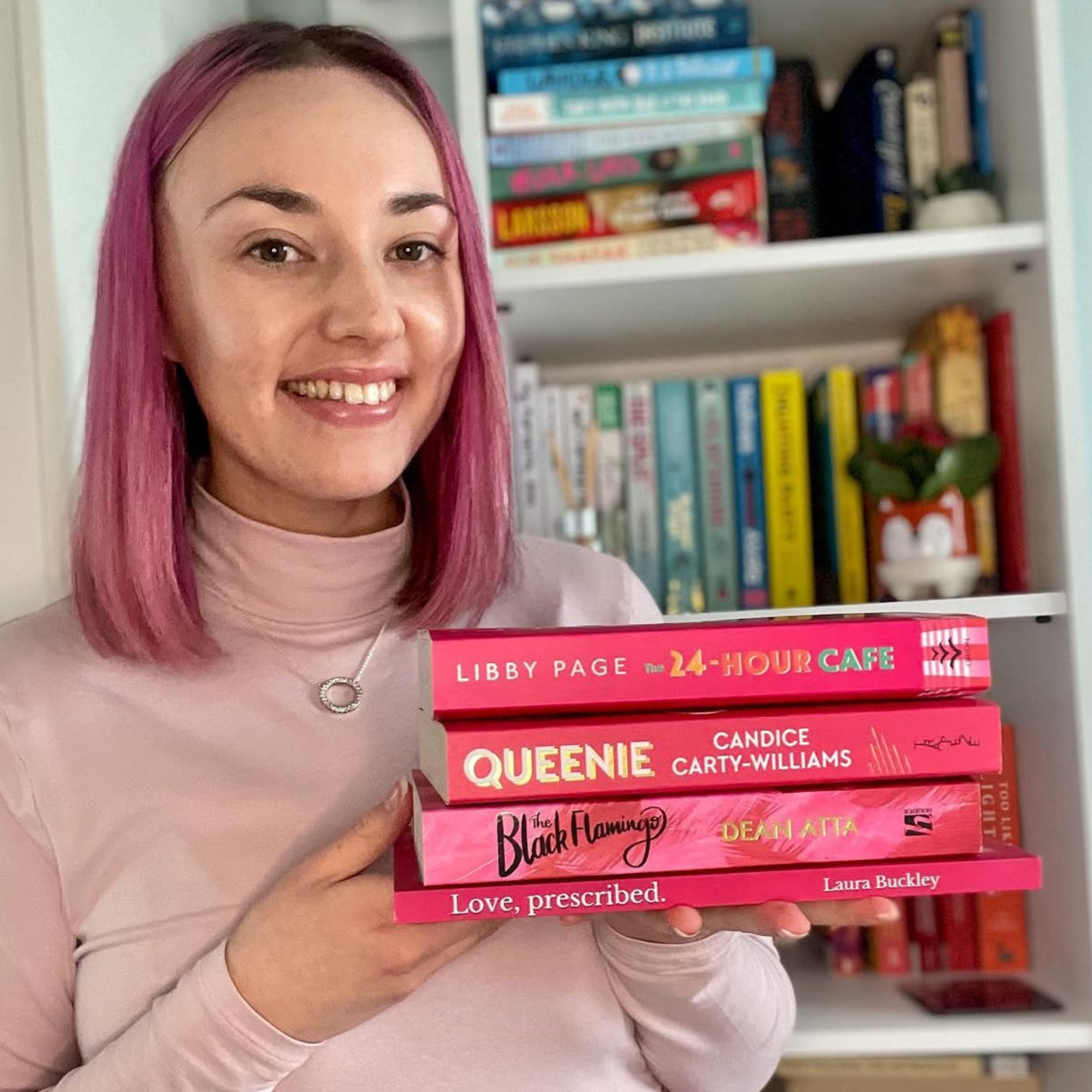 A woman with pink hair smiling and holding four books, with a bookshelf in the background.