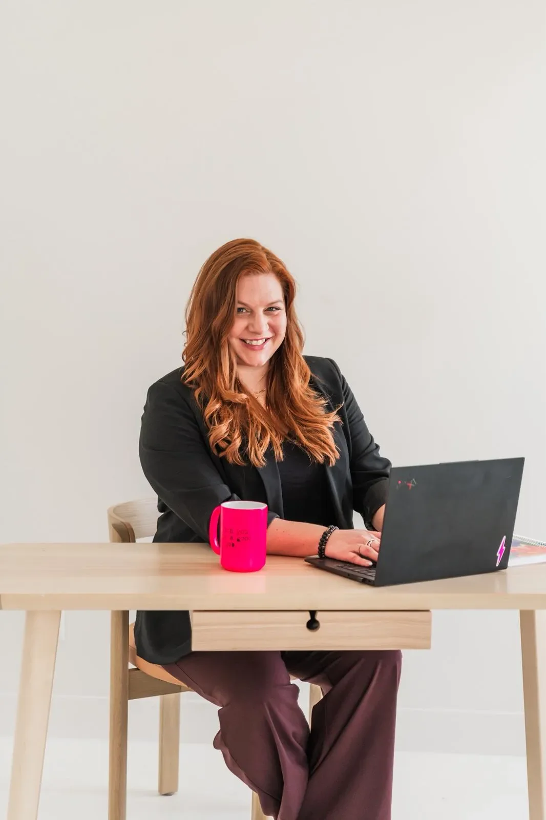 A woman with red hair smiling while sitting at a wooden table with a black laptop and a pink mug in front of her.