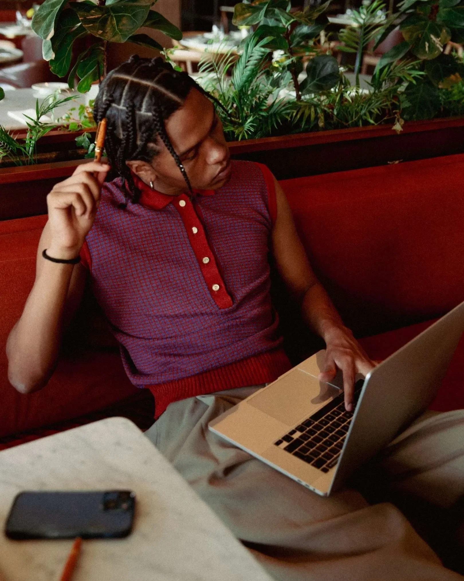 A person with braided hair sitting on a red sofa in a restaurant, using a silver laptop, with plants behind them and a smartphone on the table in front.