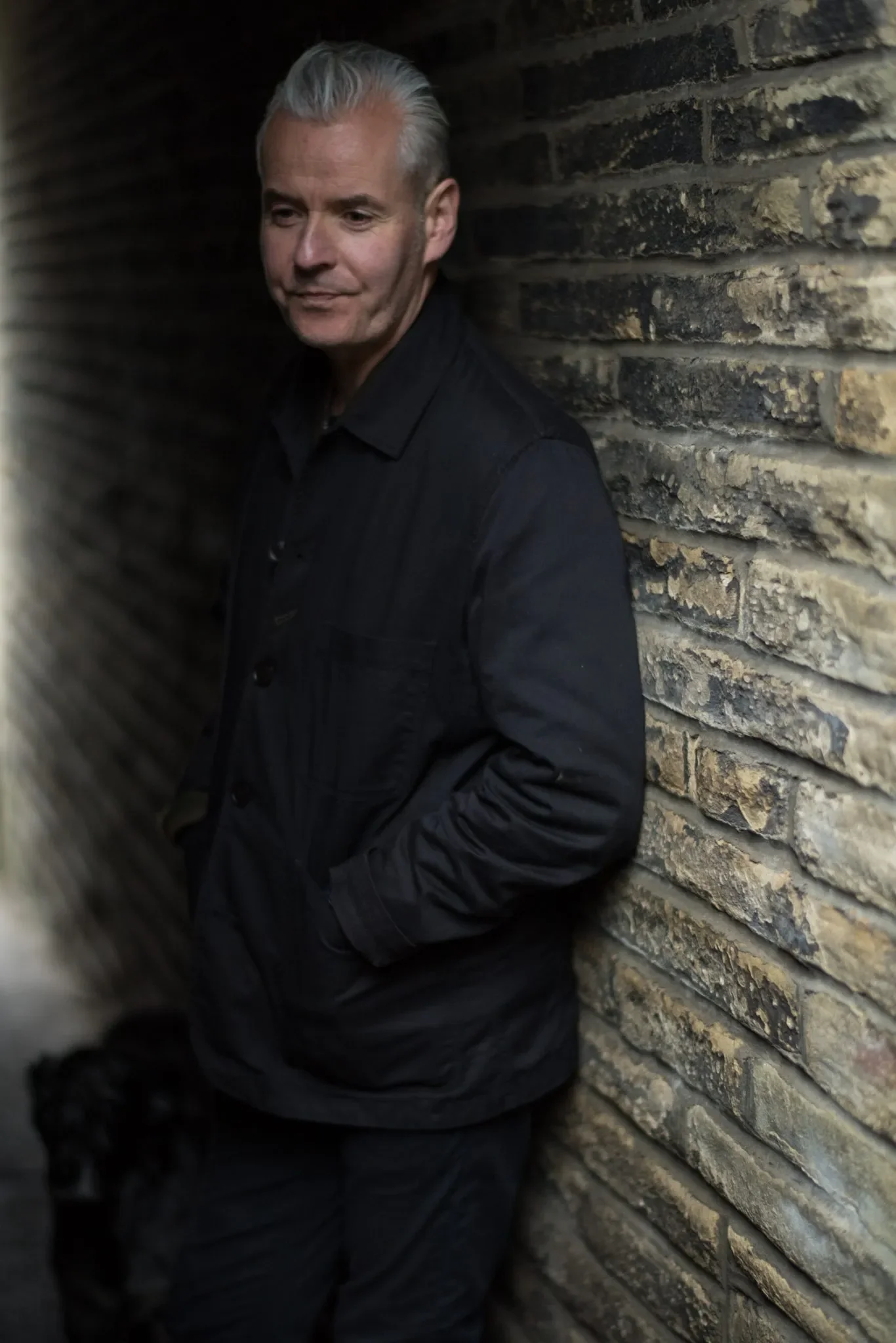 A man with gray hair and a black jacket leaning against a brick wall, with a narrow space between him and a blurry foreground.