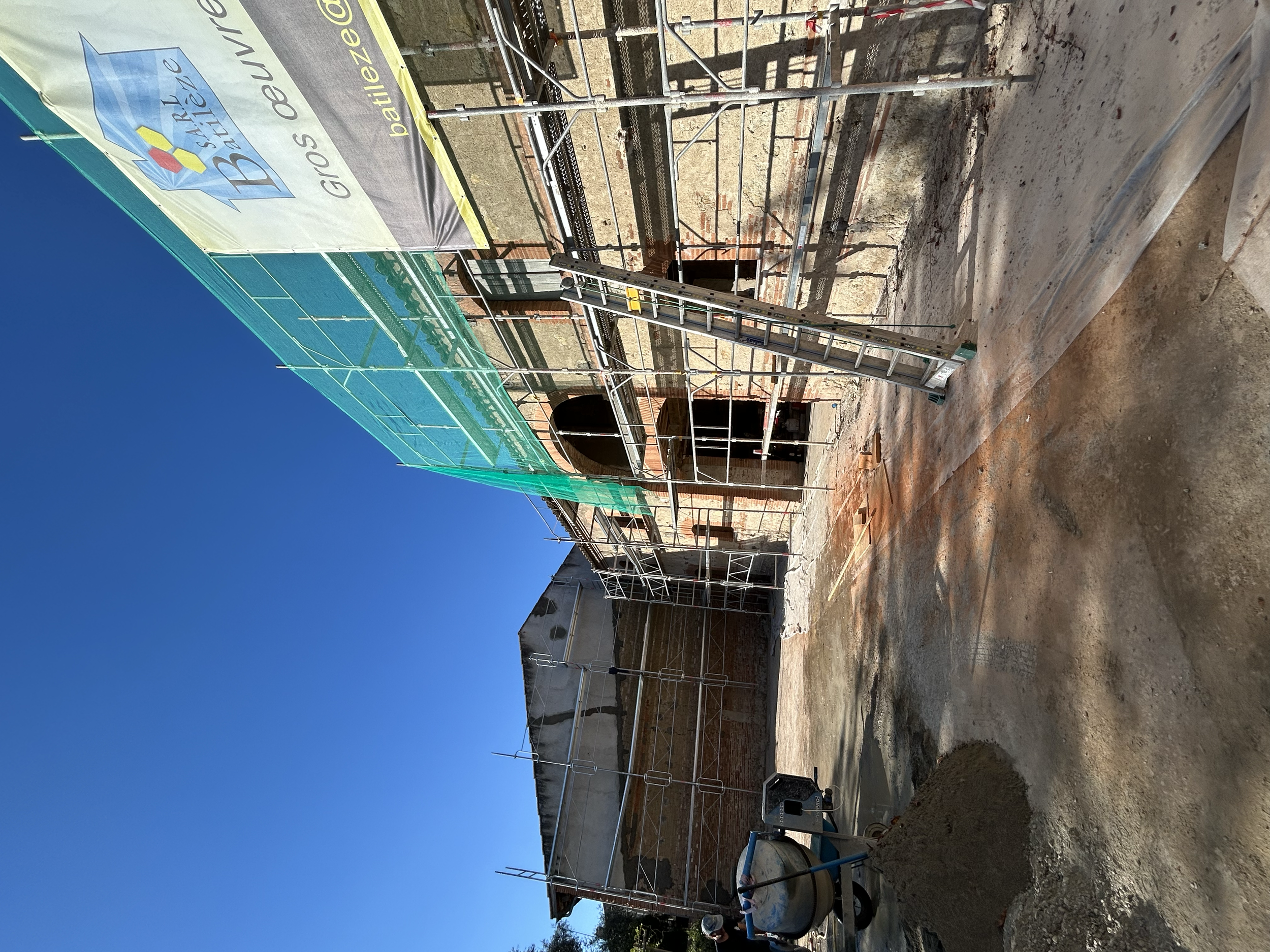 Chantier de construction avec un bâtiment en cours de rénovation, entouré d'échafaudages métalliques, sous un ciel clair. Un drapeau publicitaire est visible, ainsi qu'une truelle et un mélangeur de béton sur le sol.