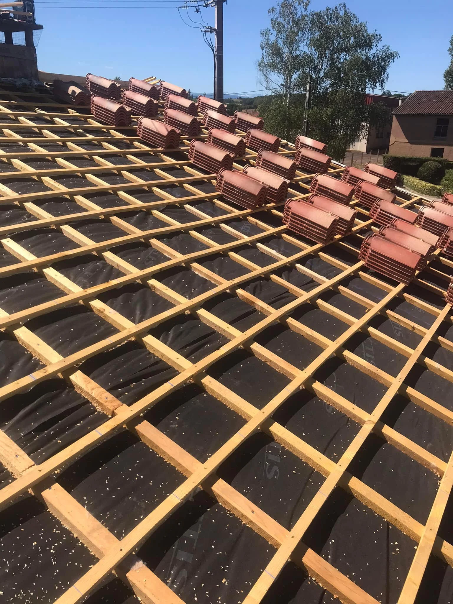 Cloison de toit en construction avec tuiles en attente d'installation, montrant une structure de bois et certaines tuiles empilées, sous un ciel ensoleillé.