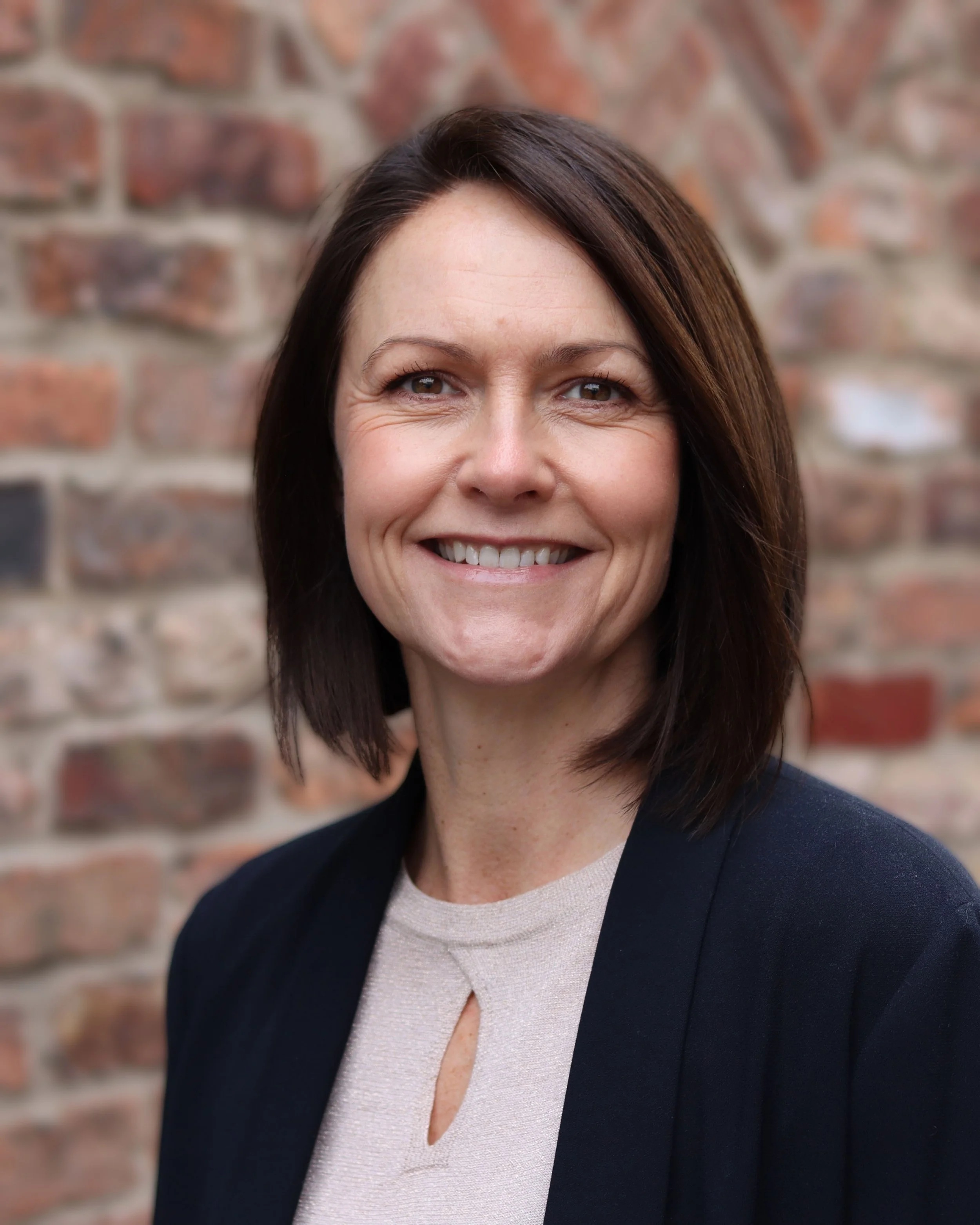 A picture of Natalie Griffin (Director) with shoulder-length dark brown hair, smiling, wearing a navy blazer and a beige top, standing against a brick wall background.