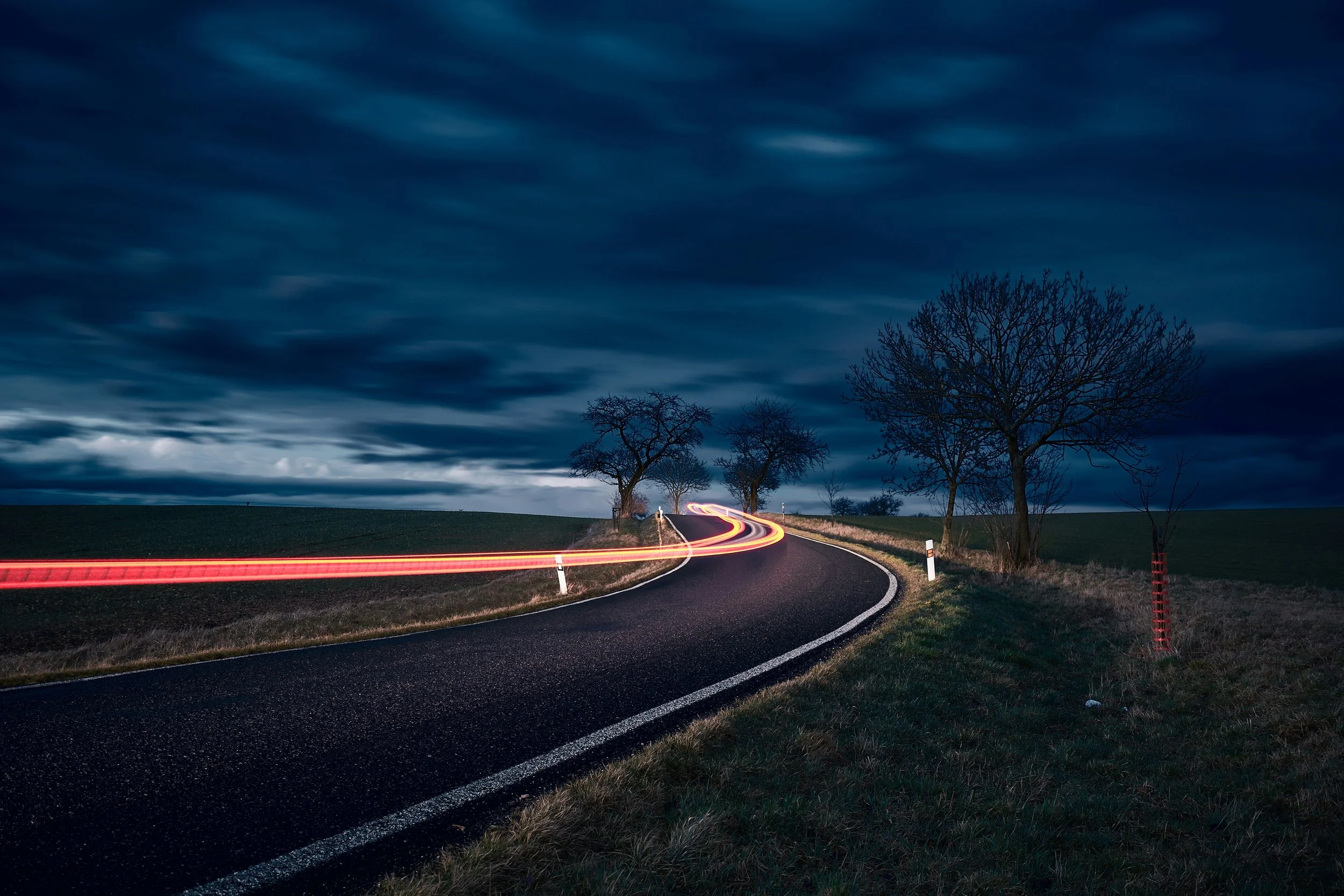 A winding country road at night with light trails from passing vehicles, bordered by grass and leafless trees under a dark, cloudy sky.