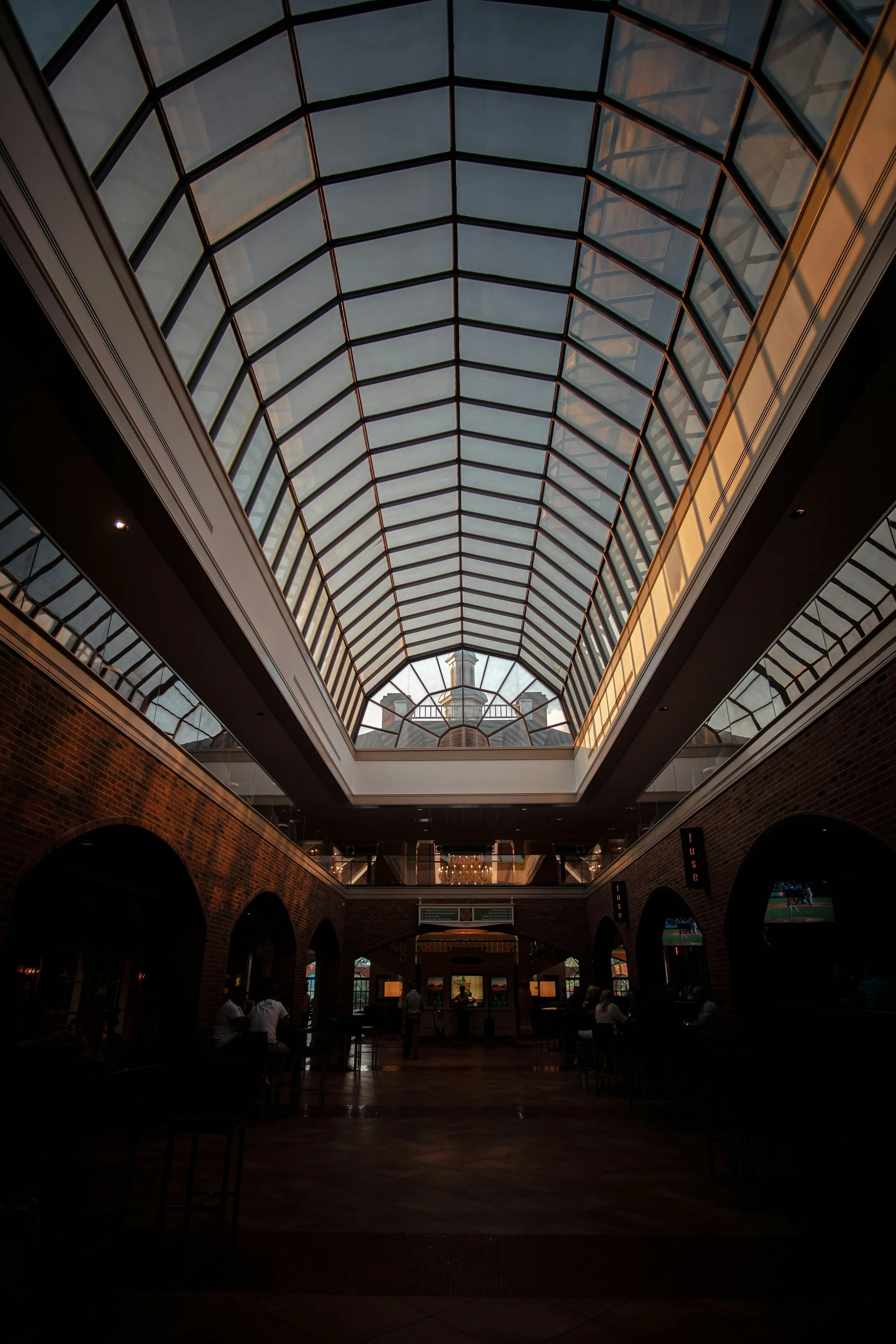 Interior view of a building with a large glass ceiling, brick walls, and seating areas with people. The ceiling has a curved, grid-like structure, allowing natural light to enter.