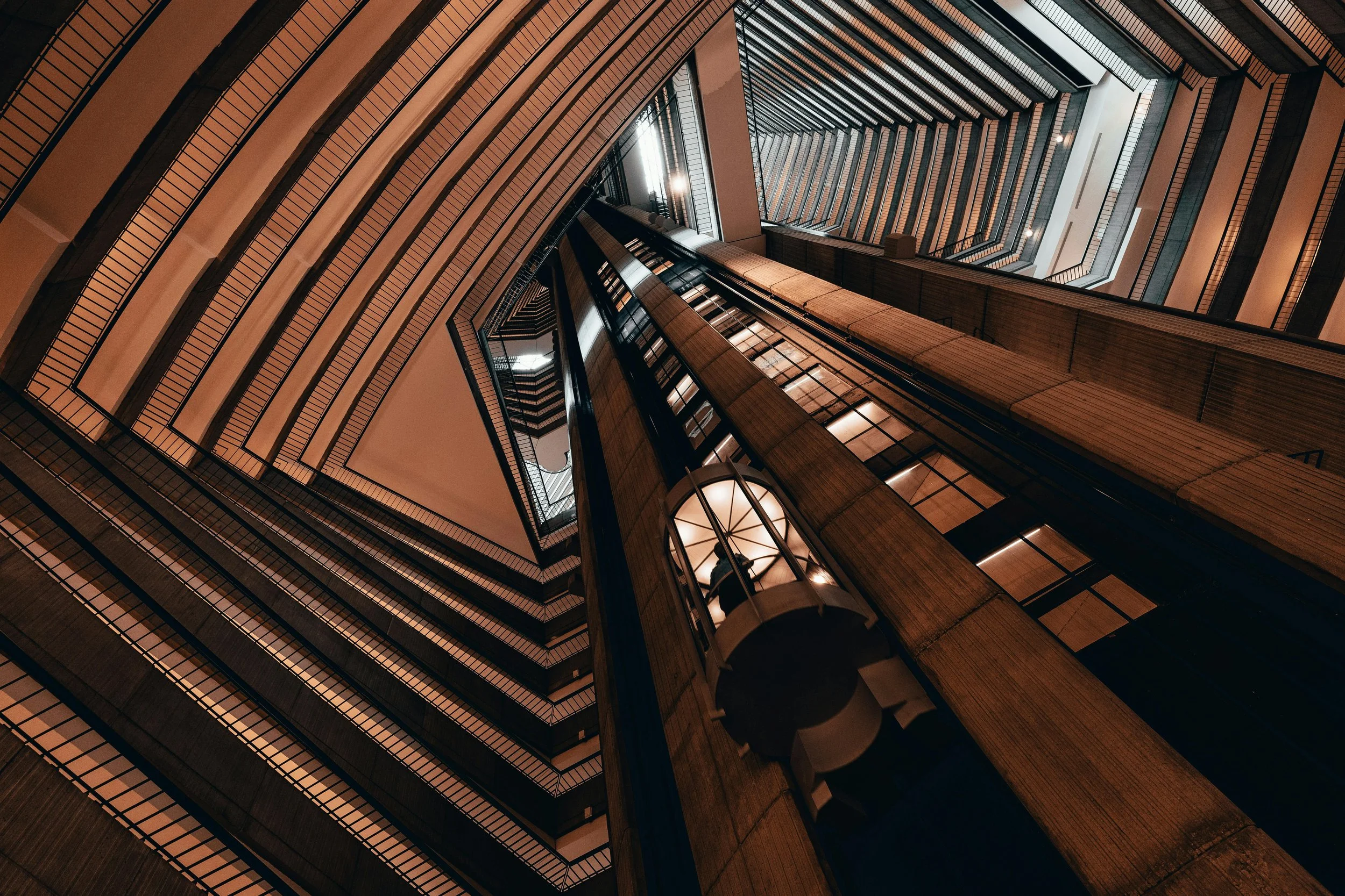 Interior view of a tall building looking upward through a central atrium with multiple floors, featuring modern architectural design with wood and glass elements, illuminated by natural and artificial lighting.