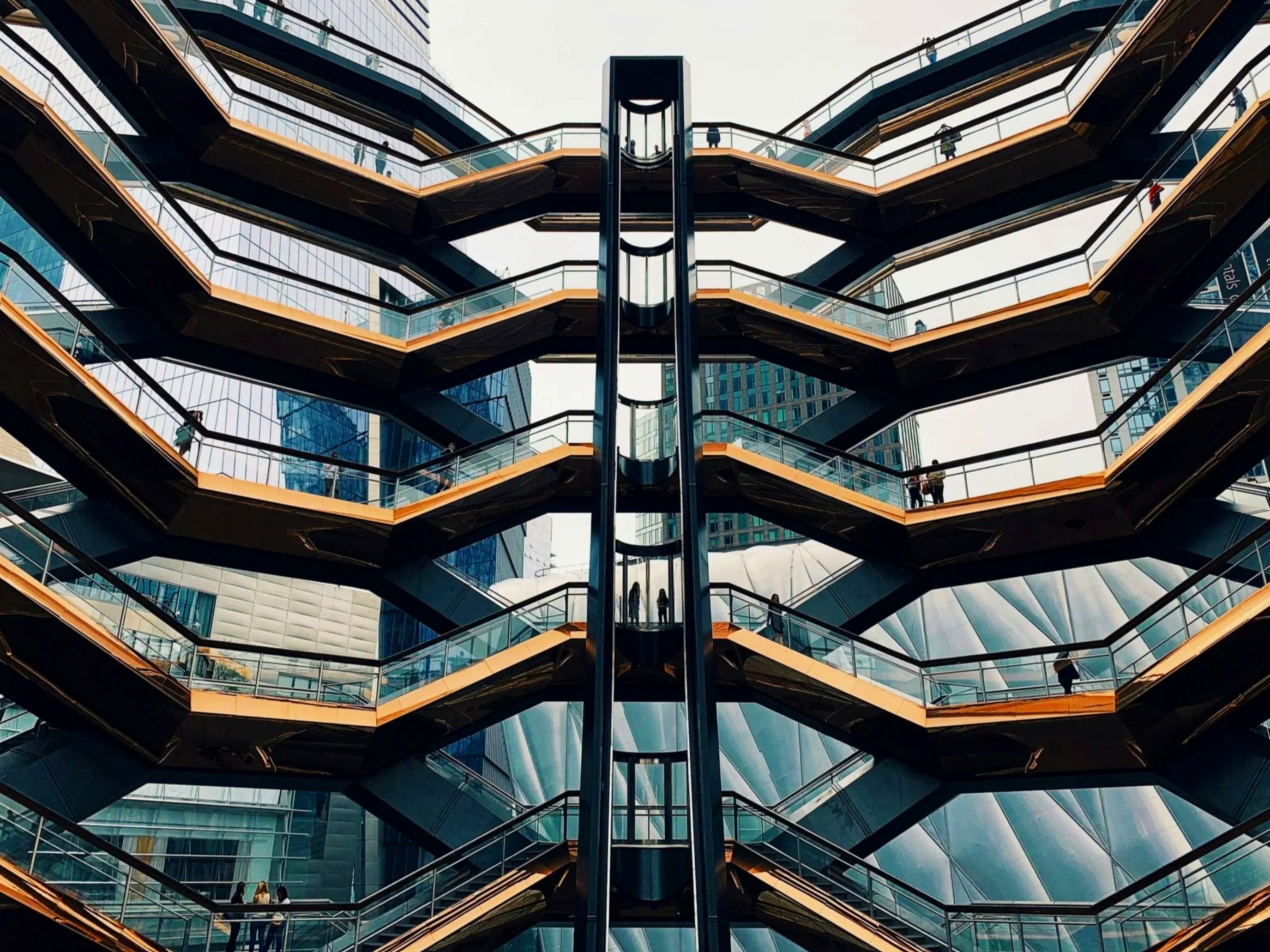 Modern multi-level outdoor escalator structure with glass and metal railings, set against tall glass buildings in an urban cityscape.