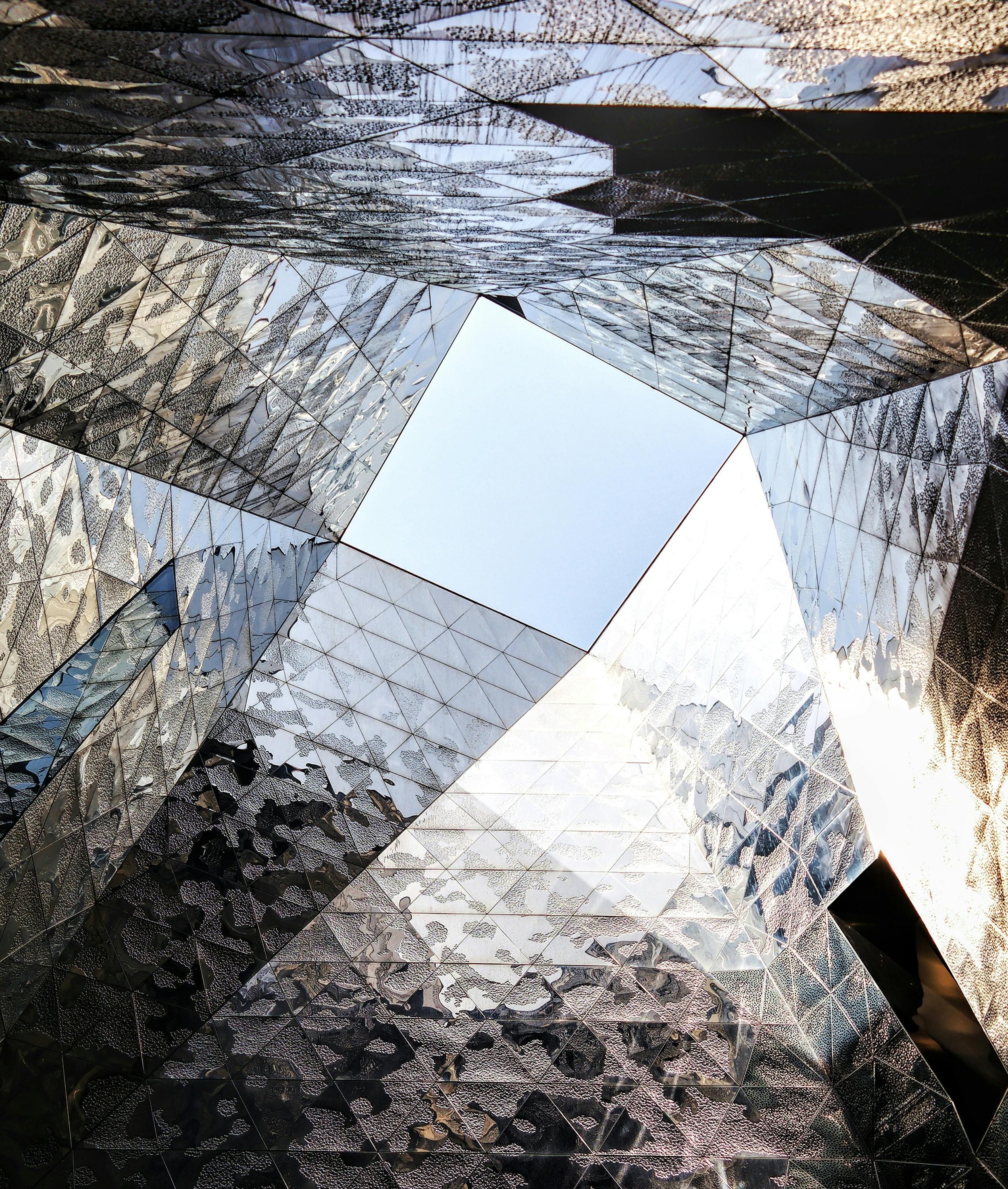 Looking up at a modern building with a square opening in the center, reflecting mirrored and textured glass panels on its exterior walls.