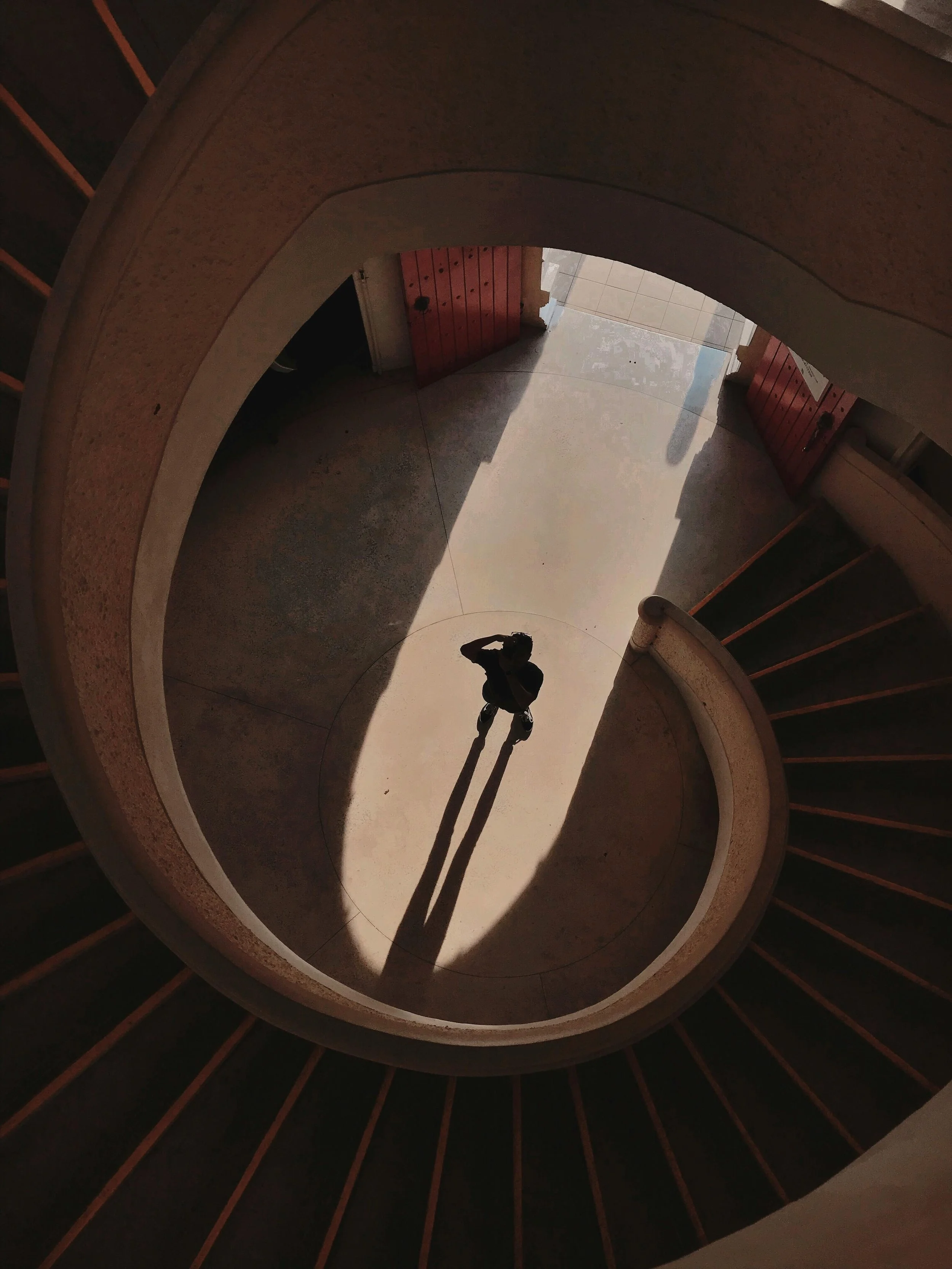 Looking down at a spiral staircase with a person taking a photo, their shadow cast on the ground, and a partially open door at the bottom of the stairs.