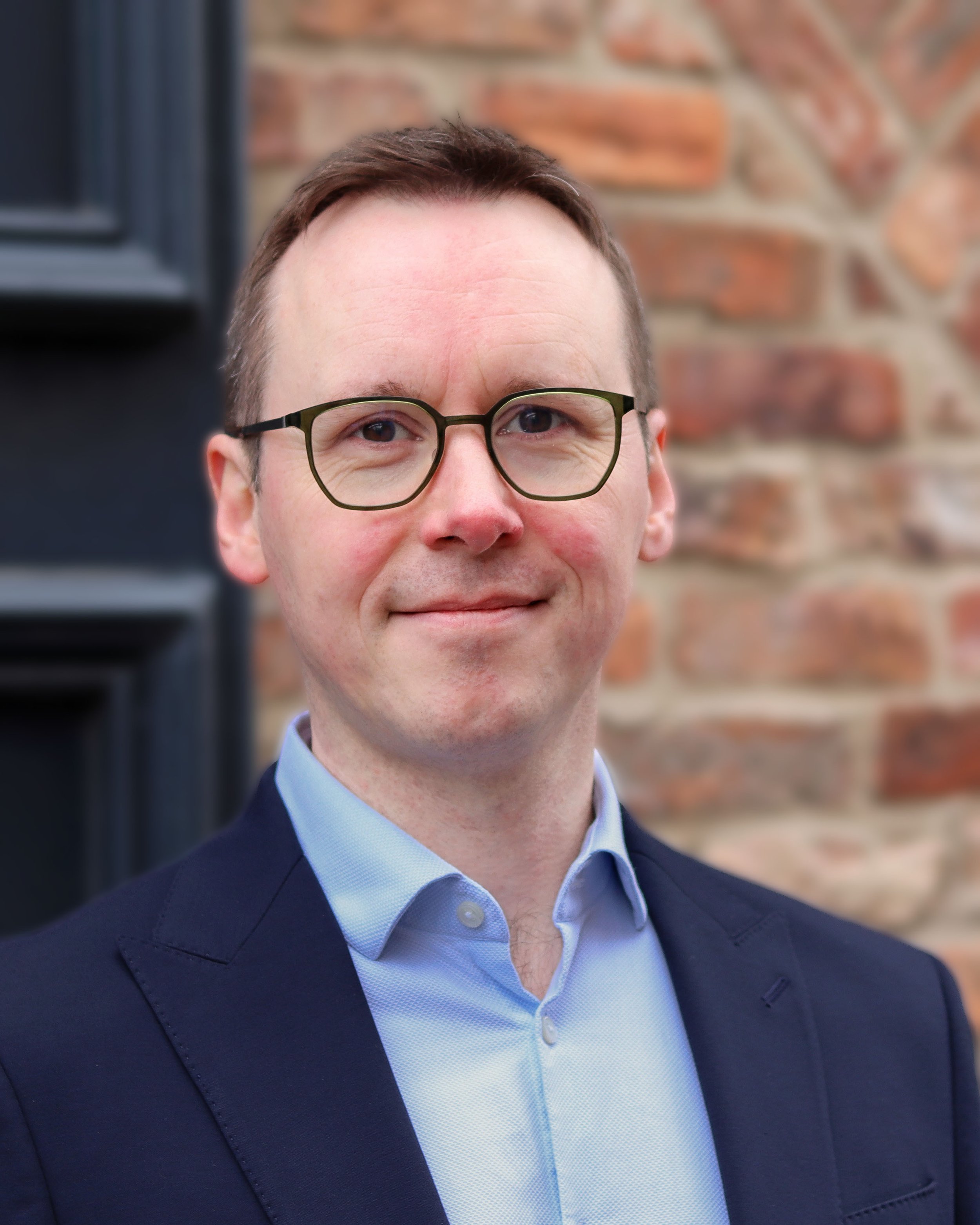 A picture of Richard Stewart (Director) wearing glasses and a suit jacket posing outdoors in front of a brick wall.