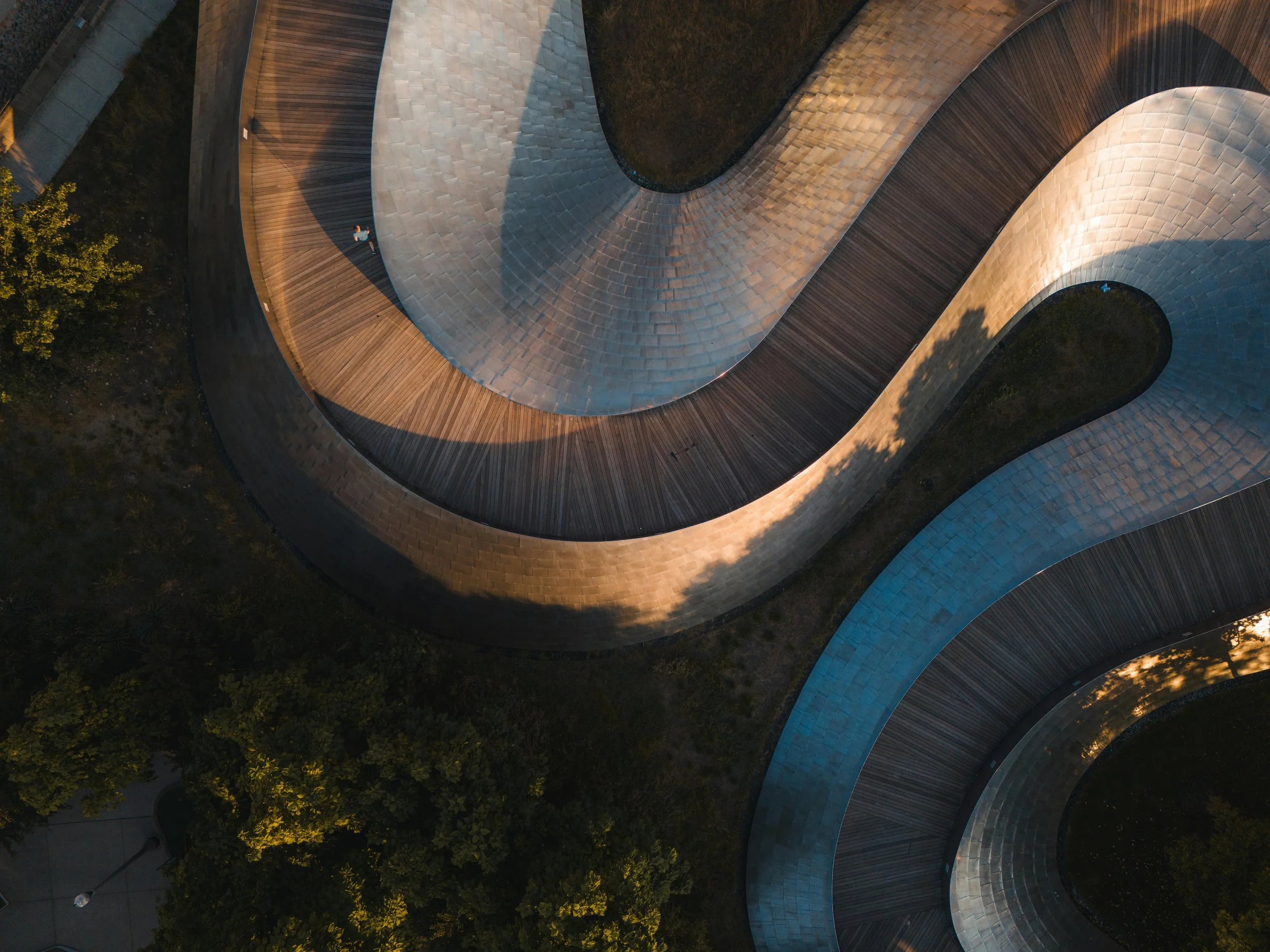 An aerial view of a modern building with a unique, curved design featuring different materials and textures, including wood and metallic panels, surrounded by greenery and trees.