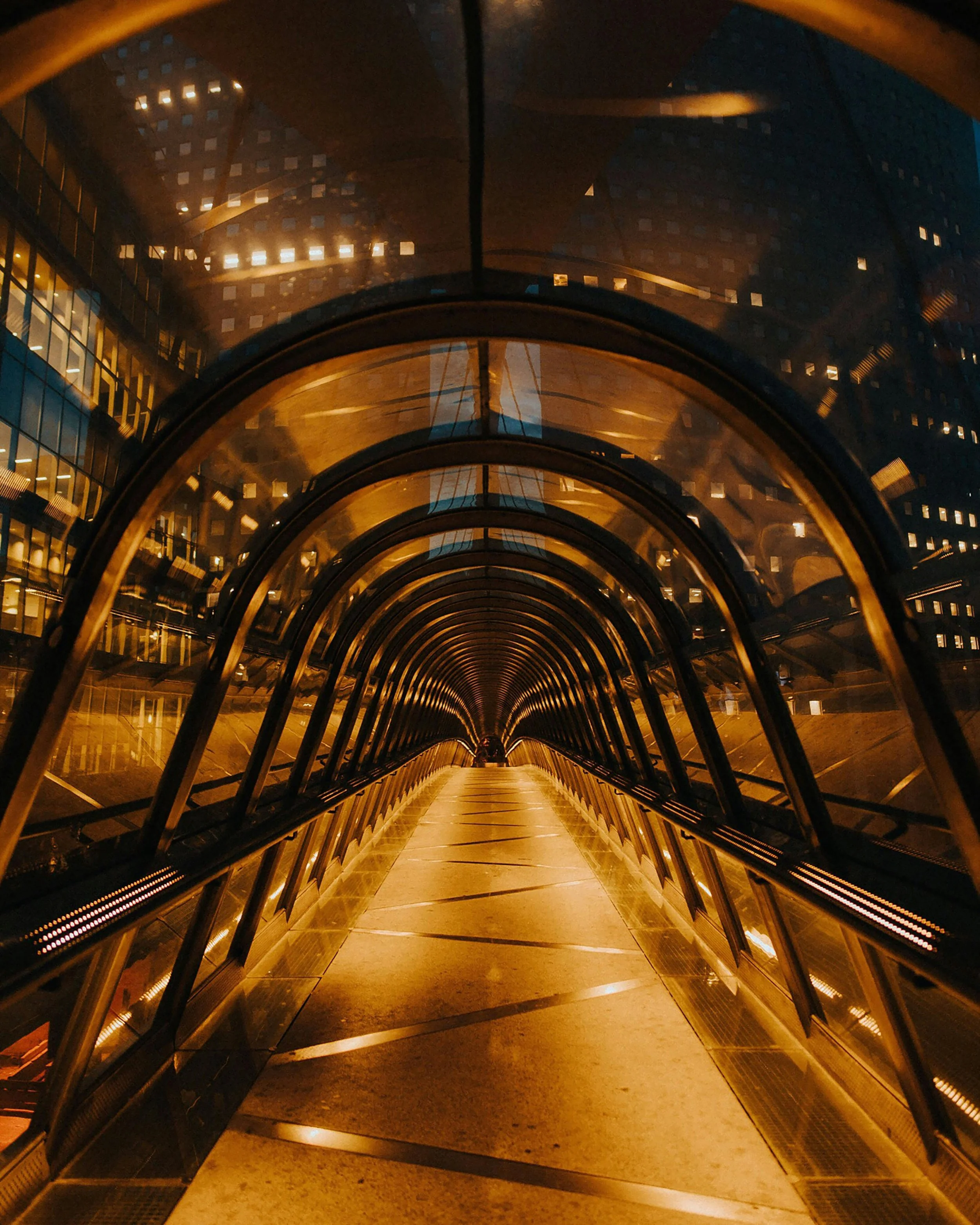A tunnel connecting two buildings at night, with a golden lighting and glass walls showing the cityscape outside.