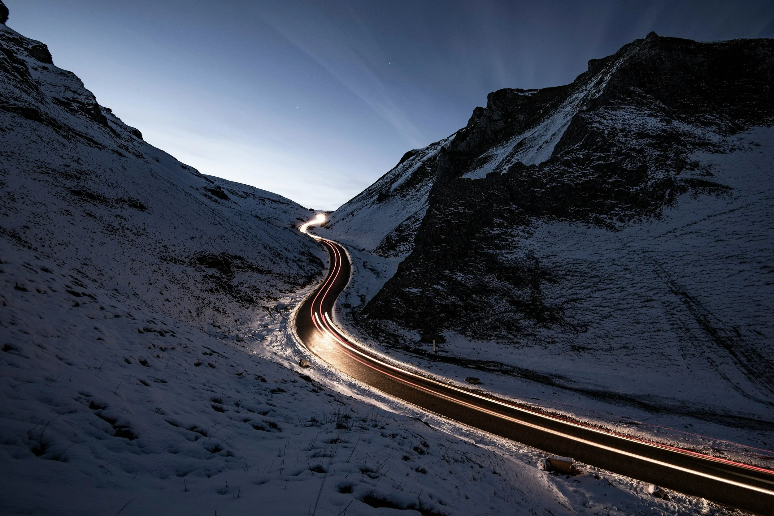 A winding mountain road at dusk with snow on the ground and light trails from moving vehicles.